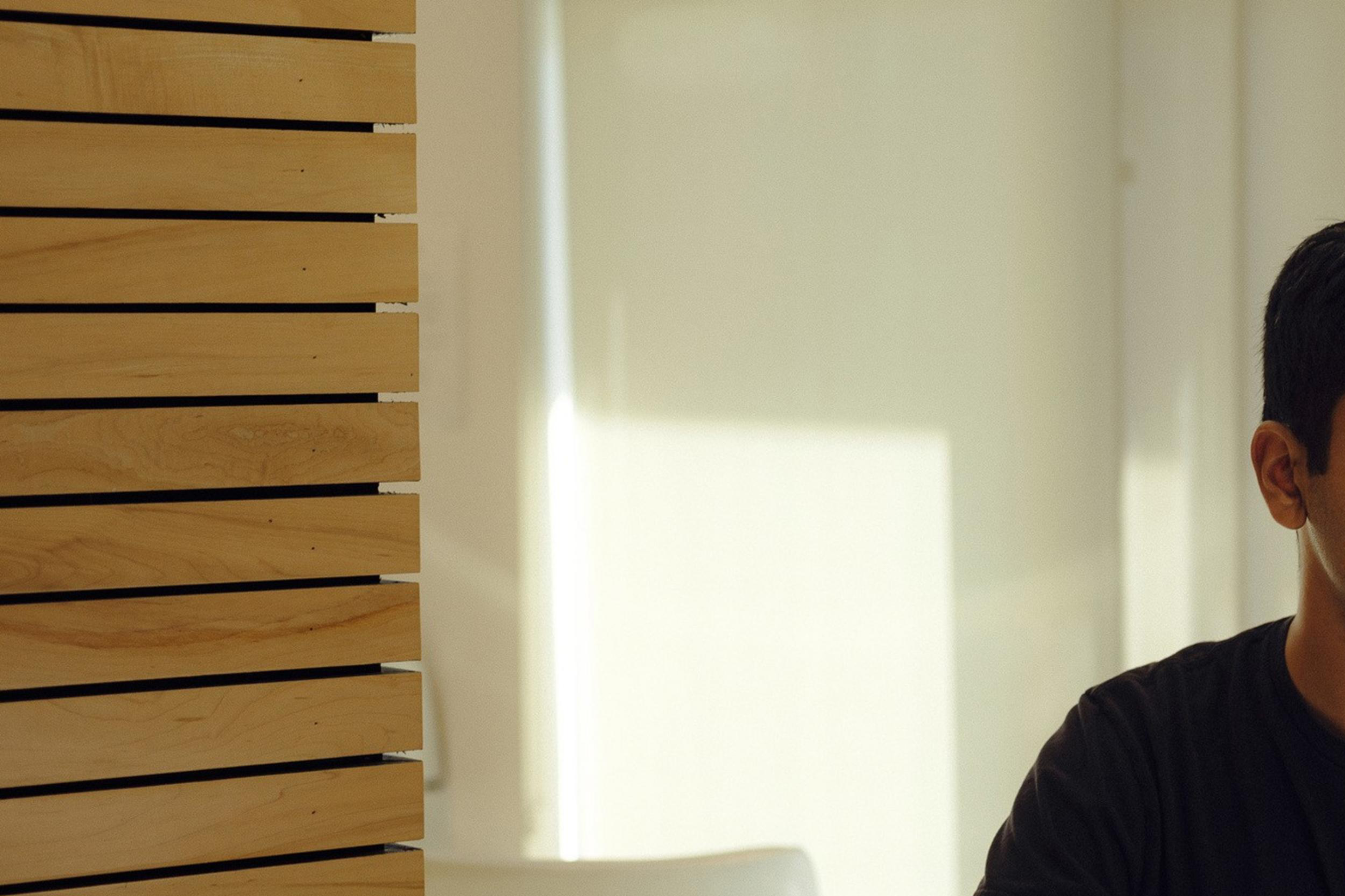 A person in a black shirt sits at a table with hands clasped. The setting is a well-lit room with a wooden panel wall and a window letting in natural light.