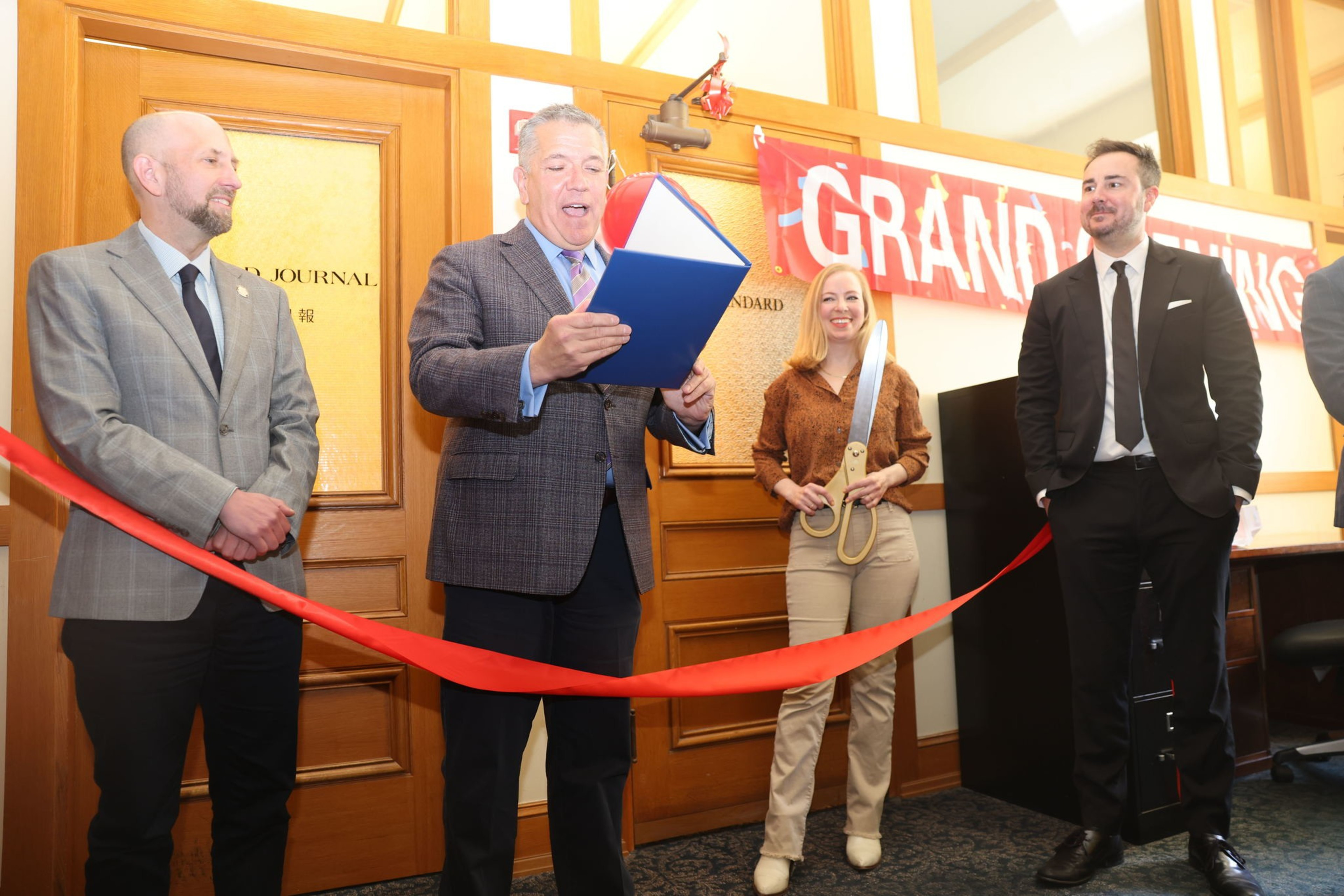 Four people stand by a red ribbon; one holds scissors, another reads from a blue folio, all smiling at a grand opening event.
