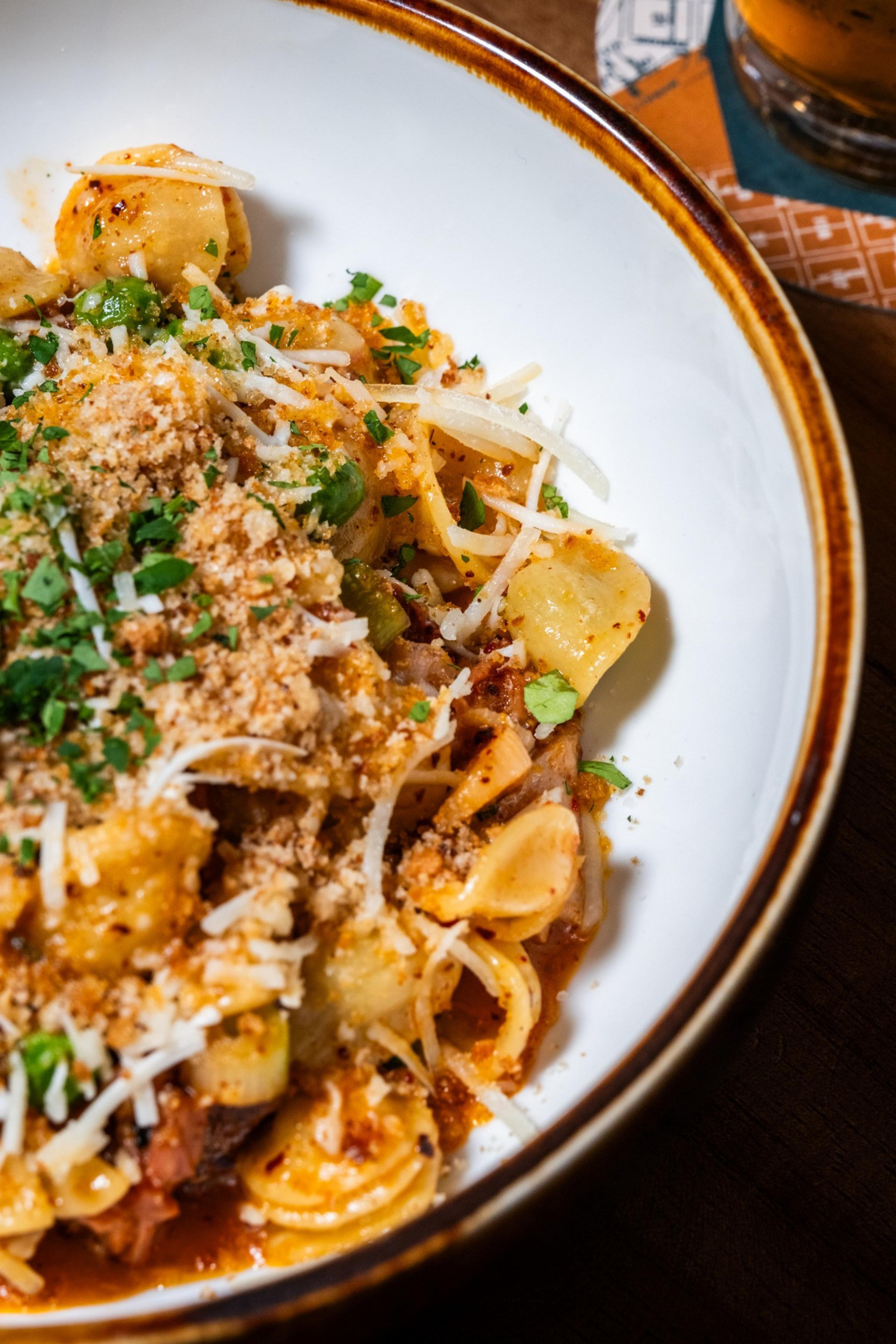 A bowl of pasta topped with grated cheese, breadcrumbs, and chopped parsley. The pasta is coated in a rich sauce with chunks of vegetables.