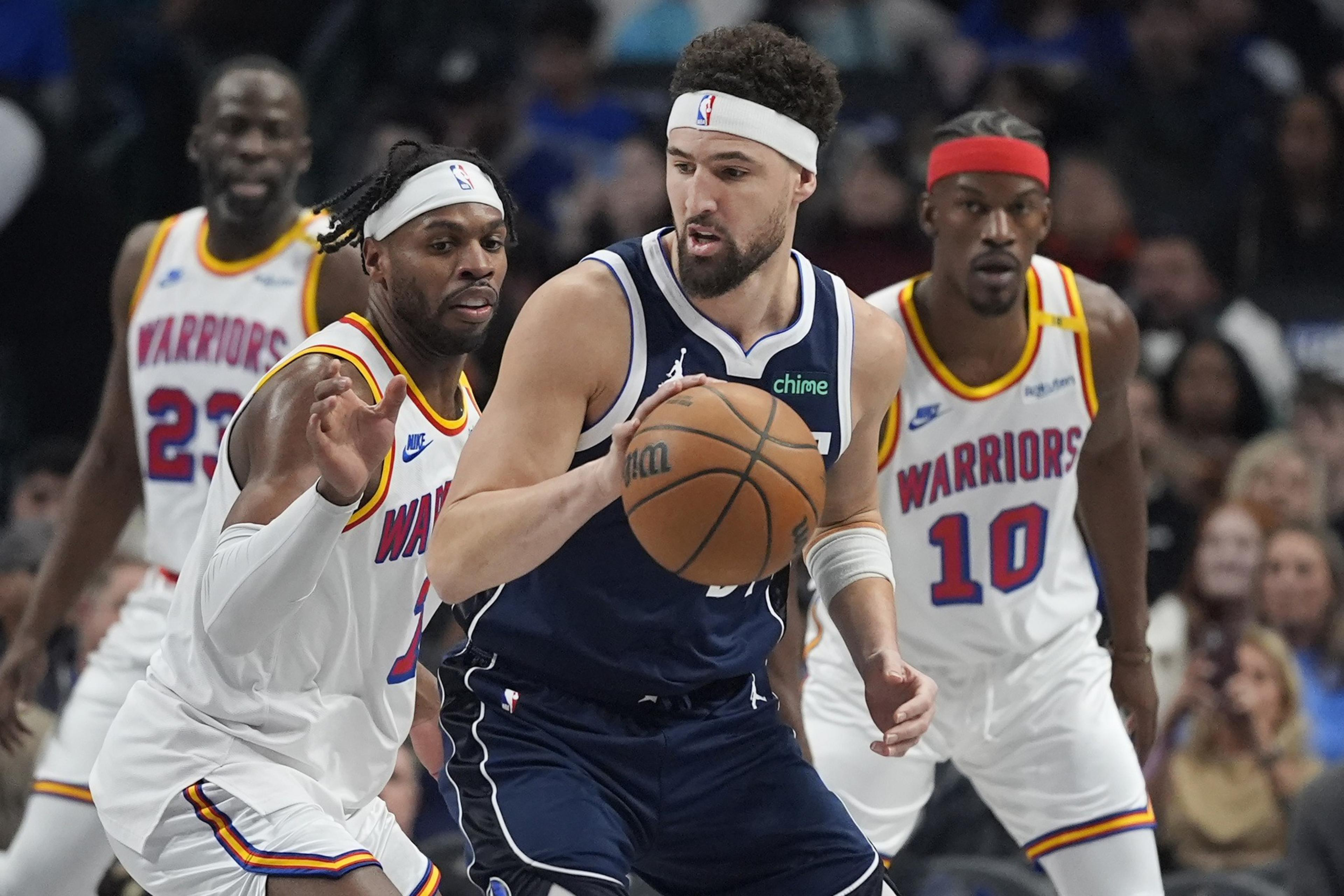 A basketball player in a blue uniform dribbles while being closely guarded by three players in white &quot;Warriors&quot; jerseys, each wearing headbands.