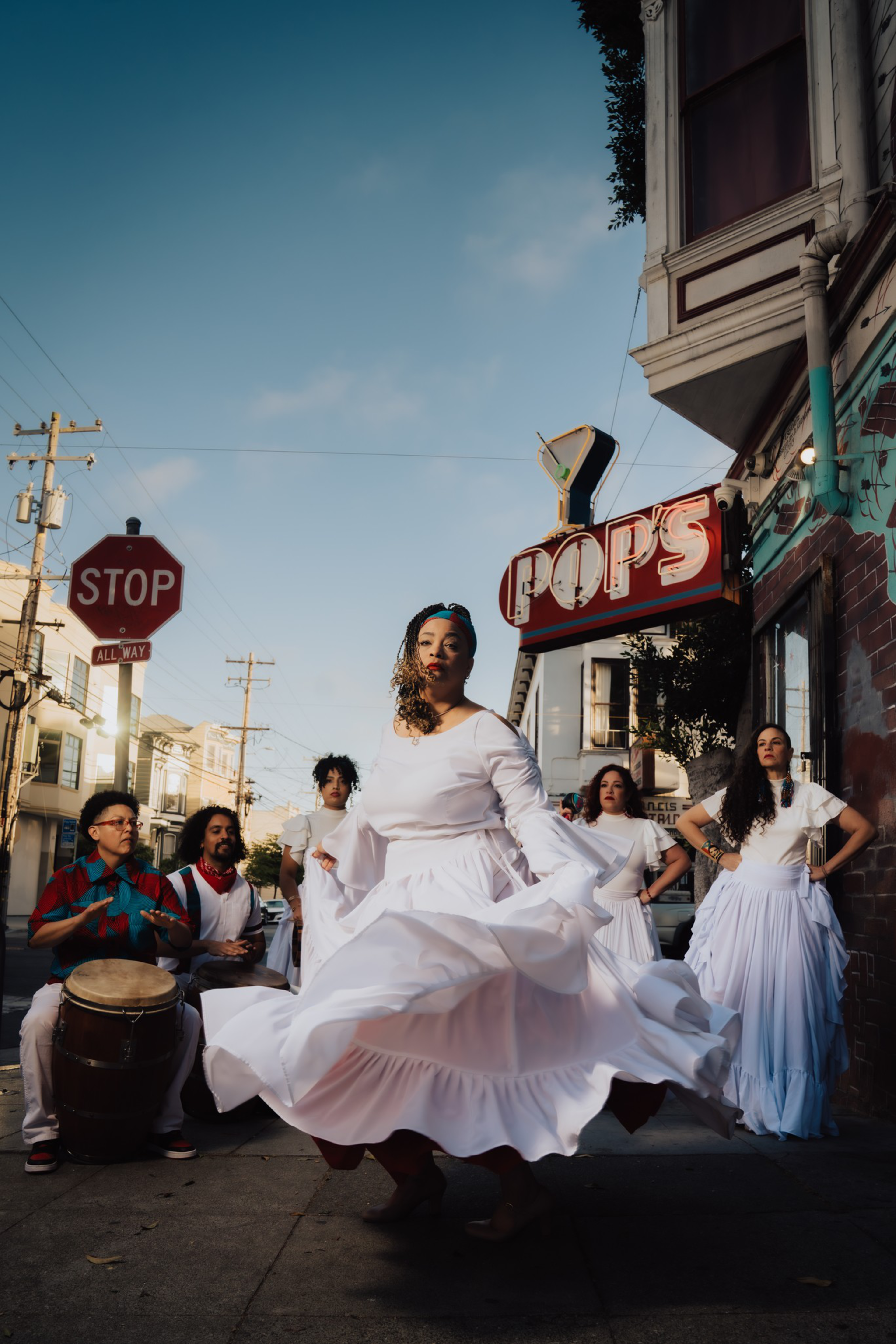 A woman in a flowing white dress spins gracefully while four women in white and two men with drums stand on a city sidewalk under a “POP’S” sign.