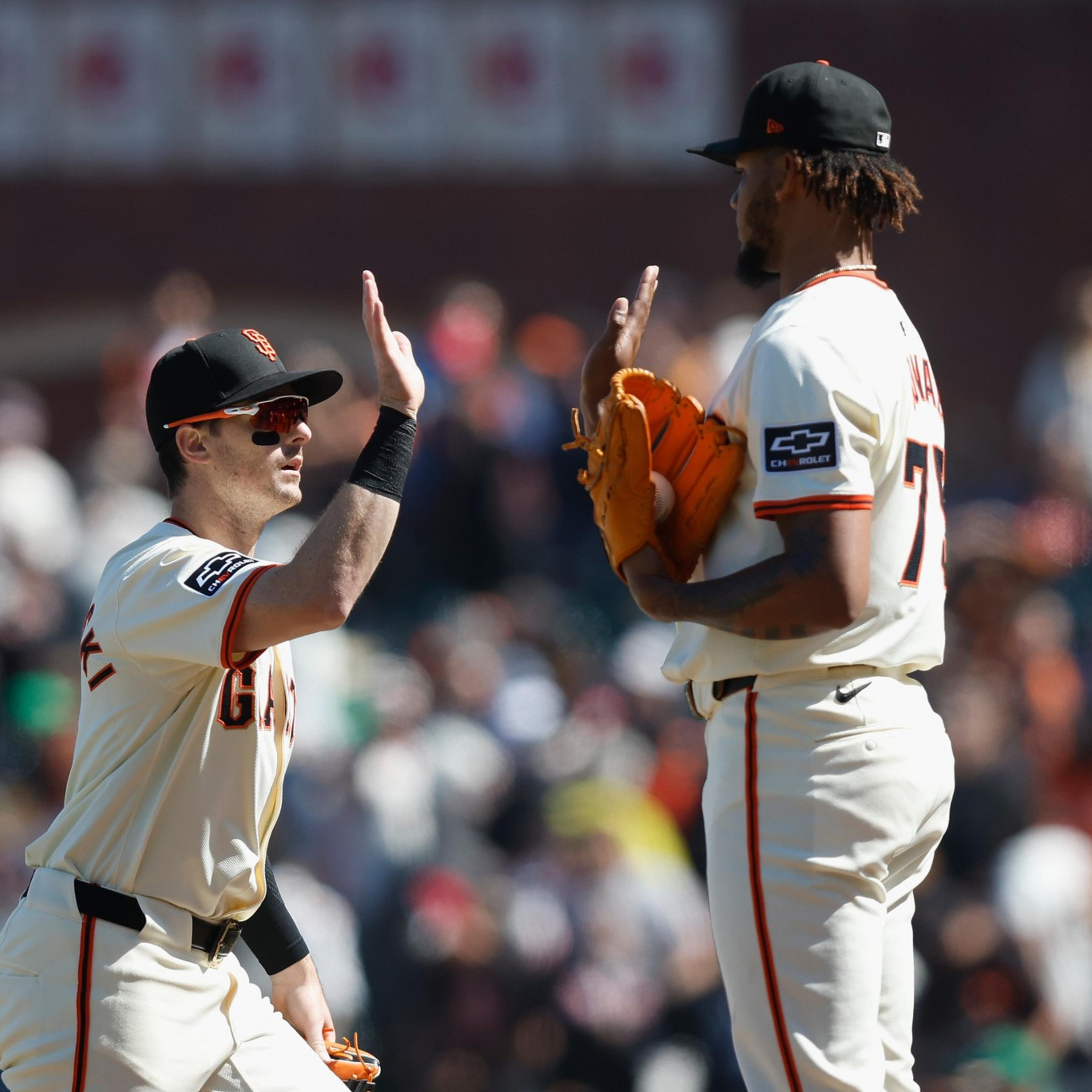 Two baseball players in white uniforms with black caps high-five each other on the field, with a blurred crowd in the background.