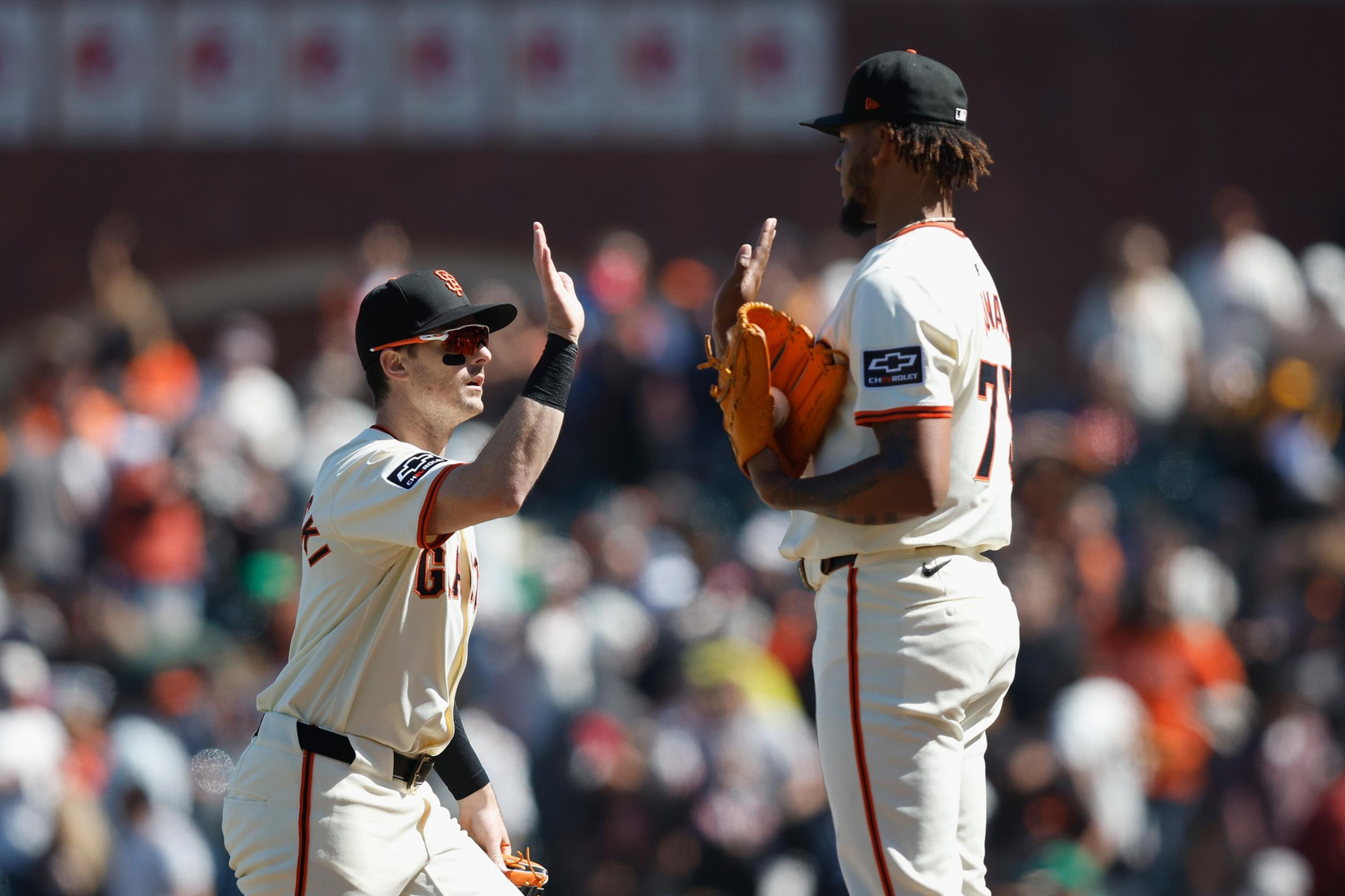 Two baseball players in white uniforms with black caps high-five each other on the field, with a blurred crowd in the background.