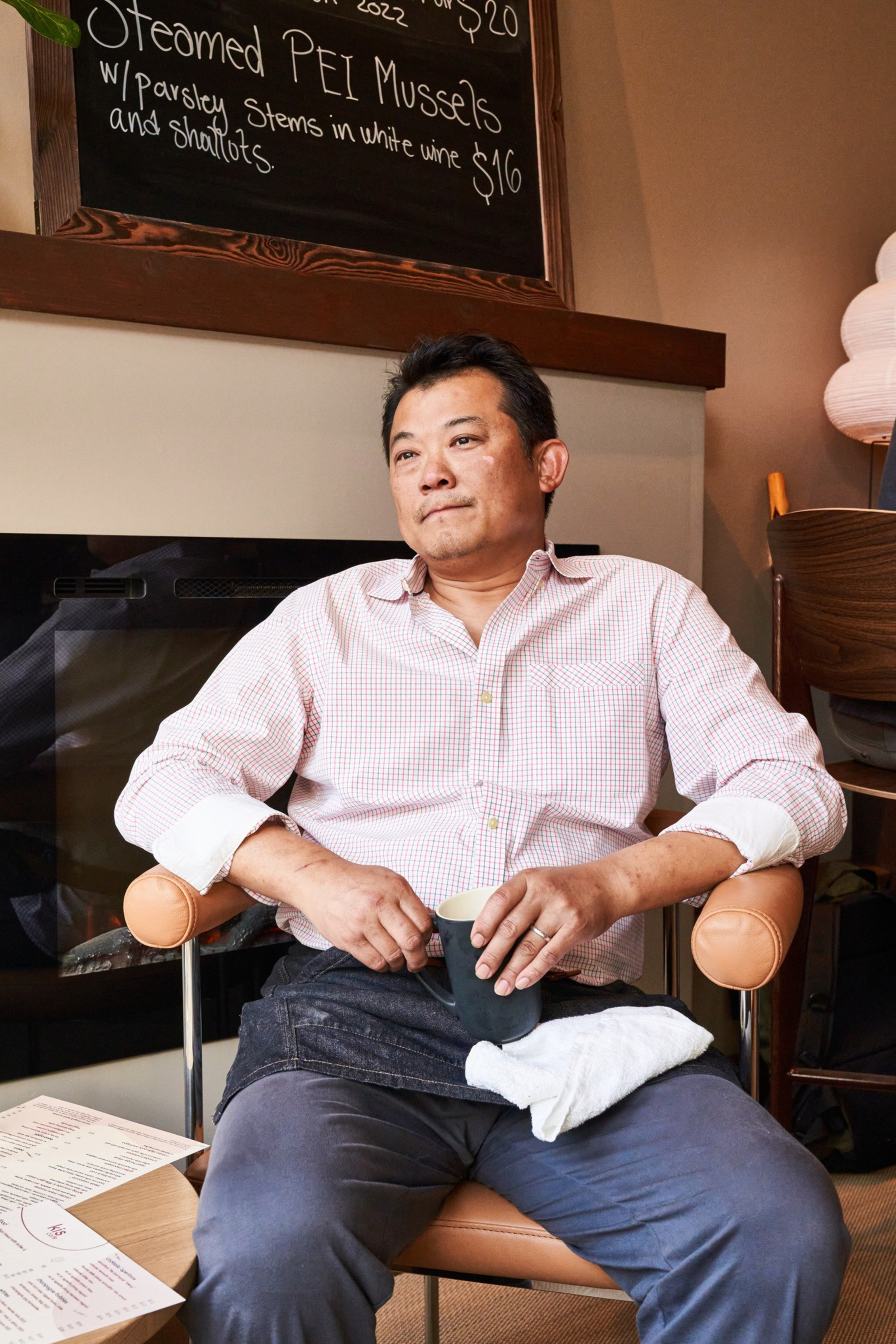 A man in a checked shirt sits in a cozy room holding a mug. Behind him, there's a chalkboard menu listing seafood items and prices.