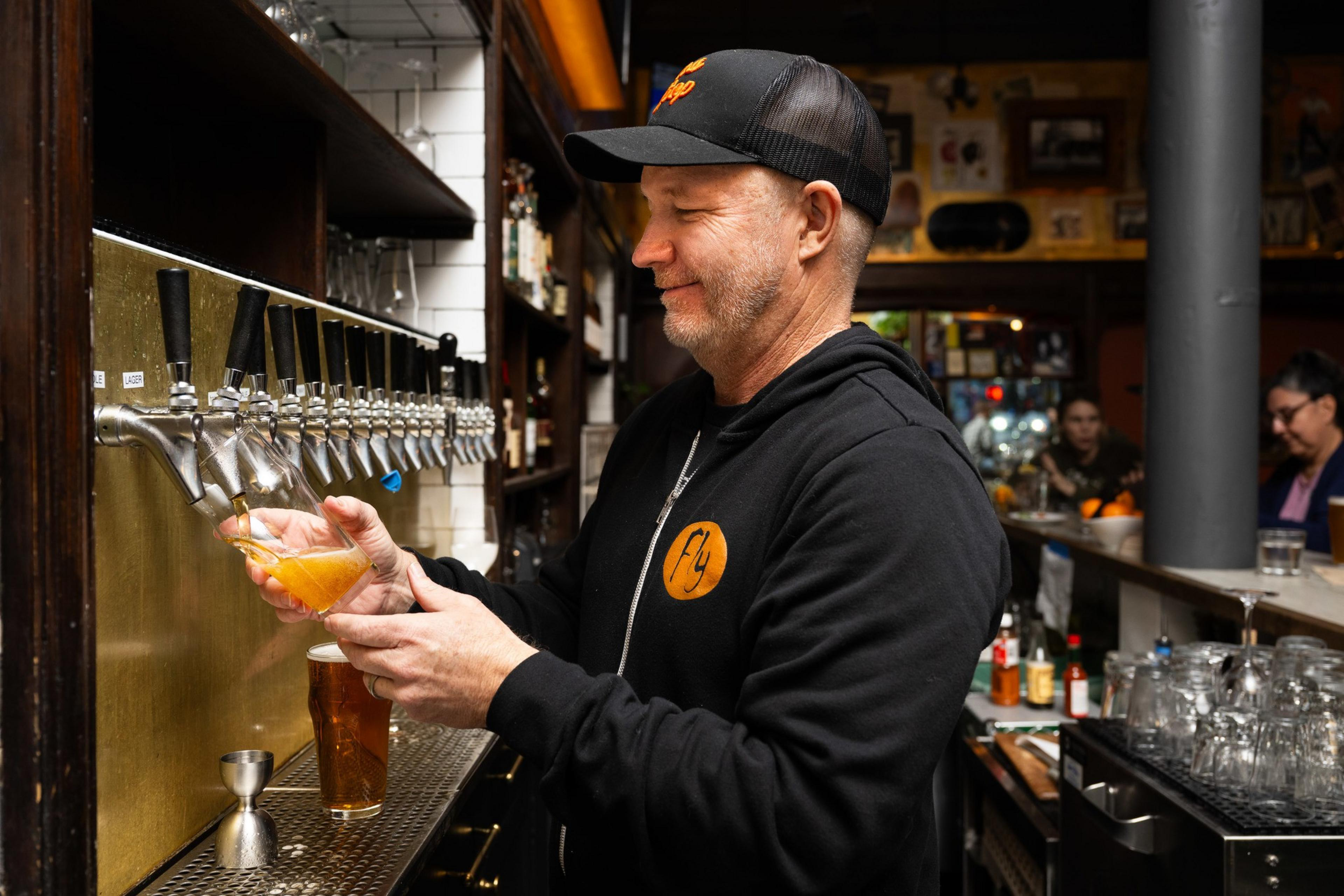 A man in a black cap and jacket pours a pint of beer from a tap in a cozy, warmly lit bar with shelves of bottles and glasses behind him.