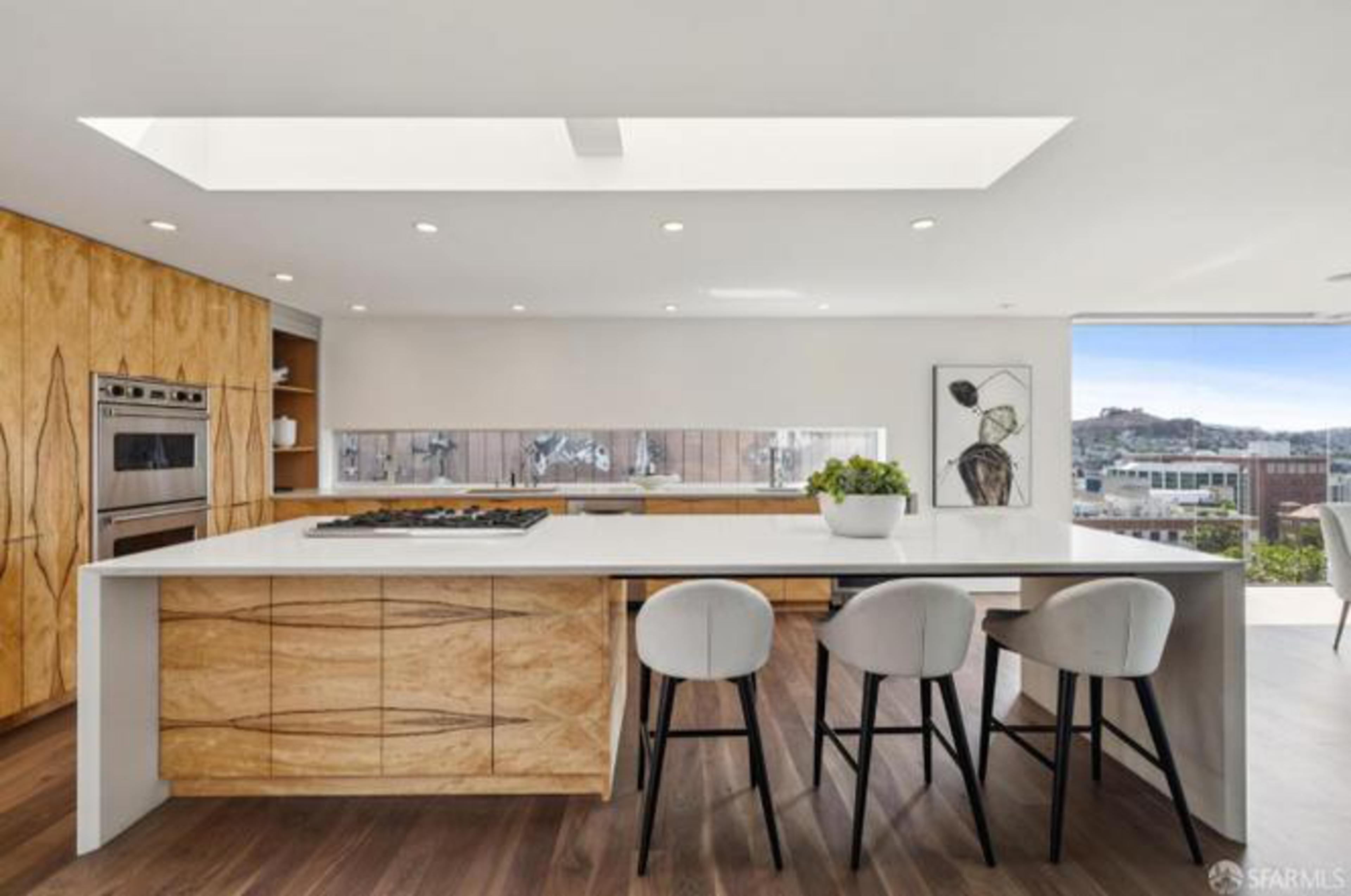 A modern kitchen with wooden cabinets, a white island countertop with three gray chairs, a built-in oven, a potted plant, and a large window showing a city view.