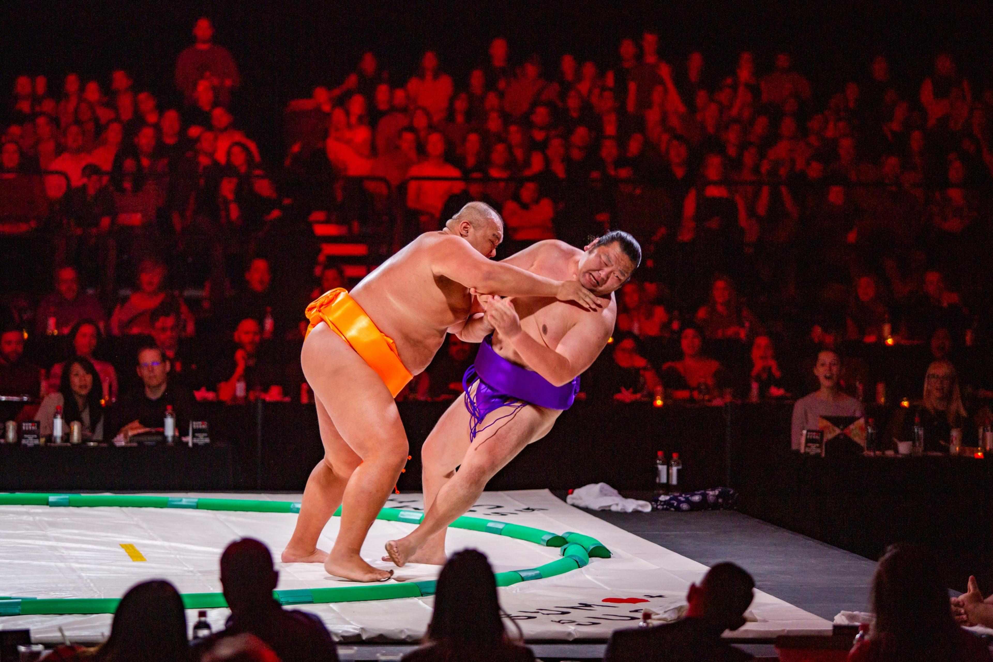 Two sumo wrestlers, one in an orange mawashi and the other in purple, grapple intensely in a ring as a captivated crowd watches in the background.