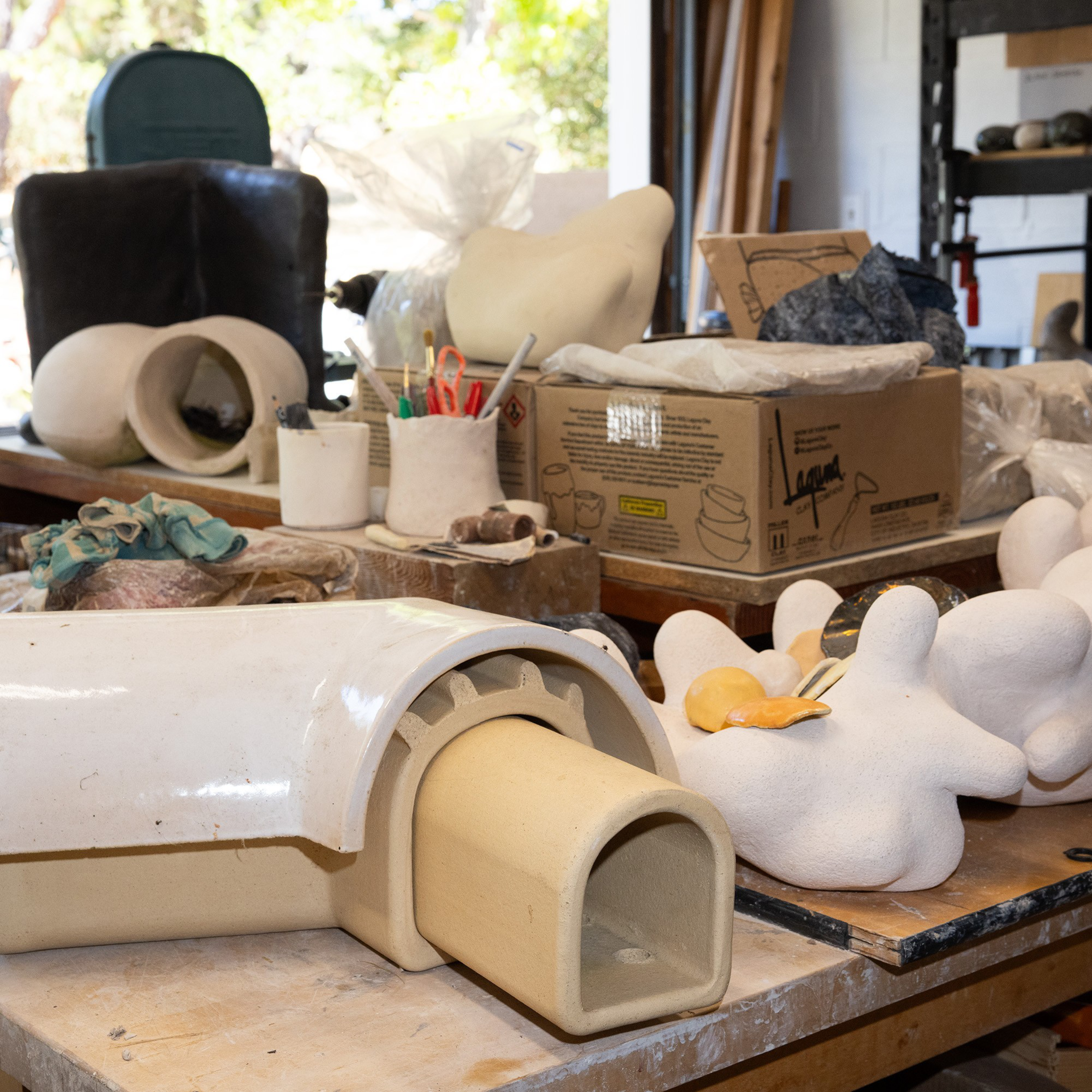 The image shows a cluttered workshop table filled with various ceramics, unfinished clay sculptures, and art supplies in containers. Shelves and a green chair are in the background.