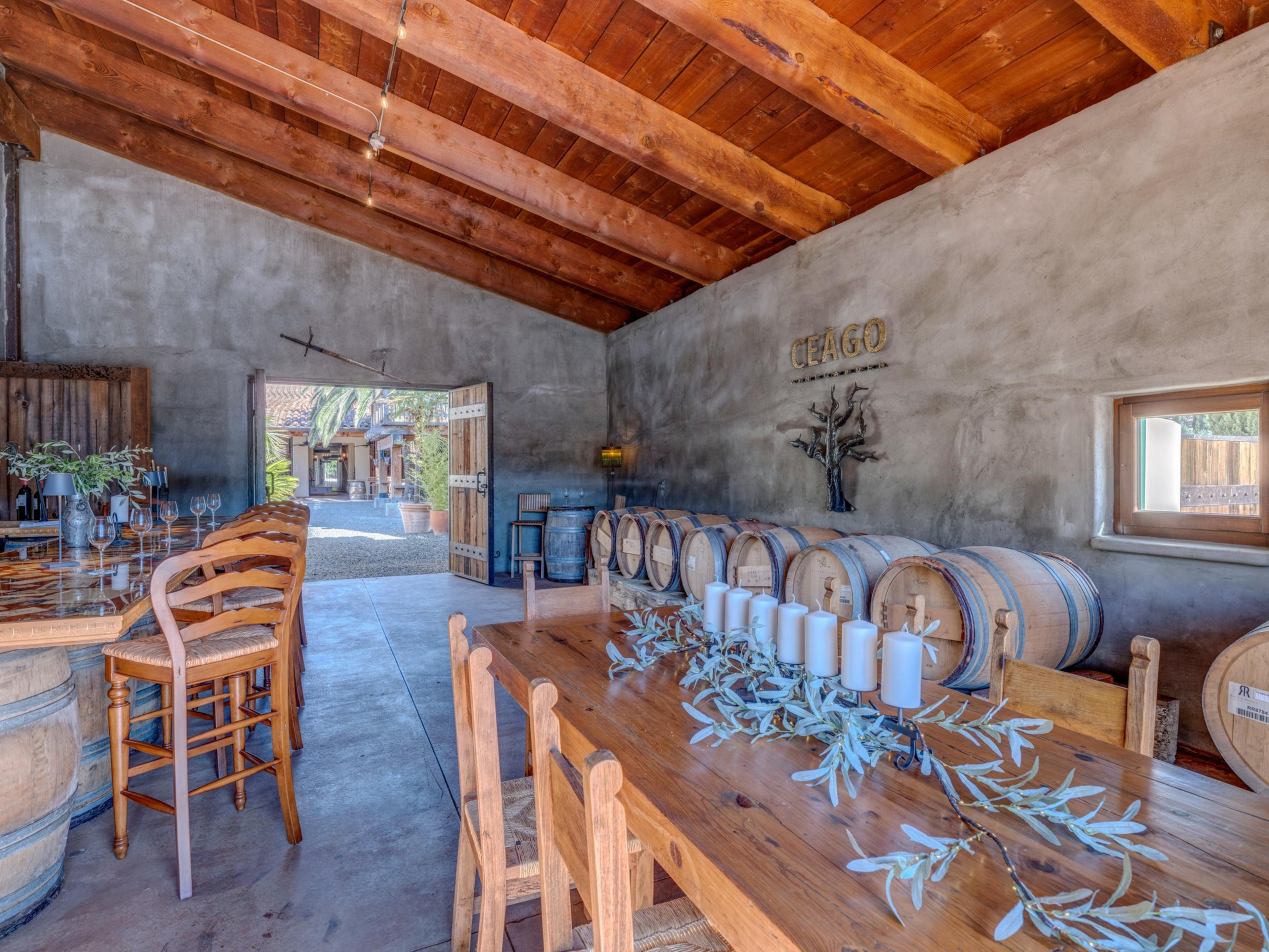 A rustic room with wooden barrels, two wooden tables with chairs, a row of white candles on one table, and an open door showing an outdoor area.