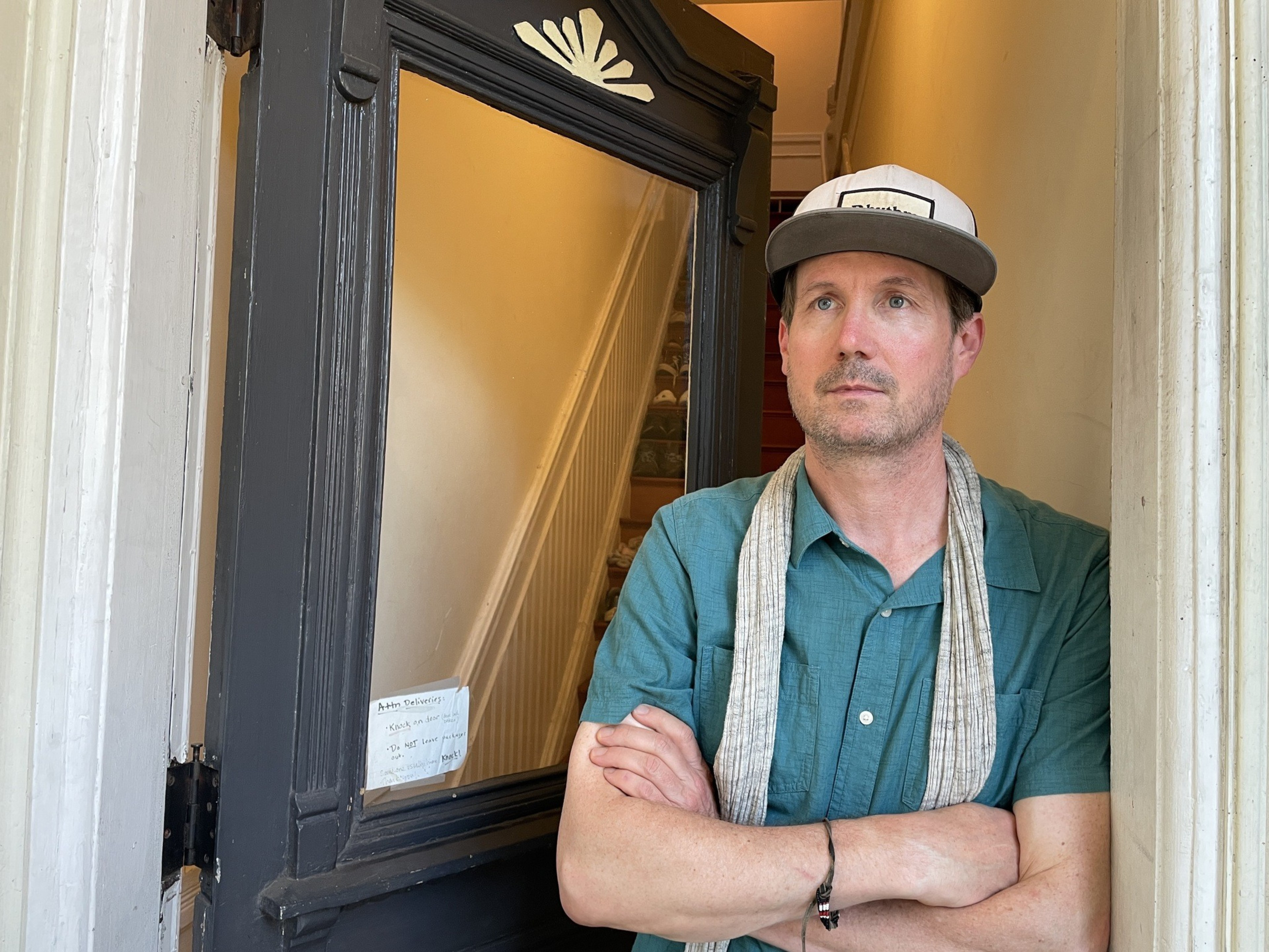 A man in a blue shirt and hat stands in a doorway with crossed arms, a beige scarf around his neck, and an interior stairway visible behind him.
