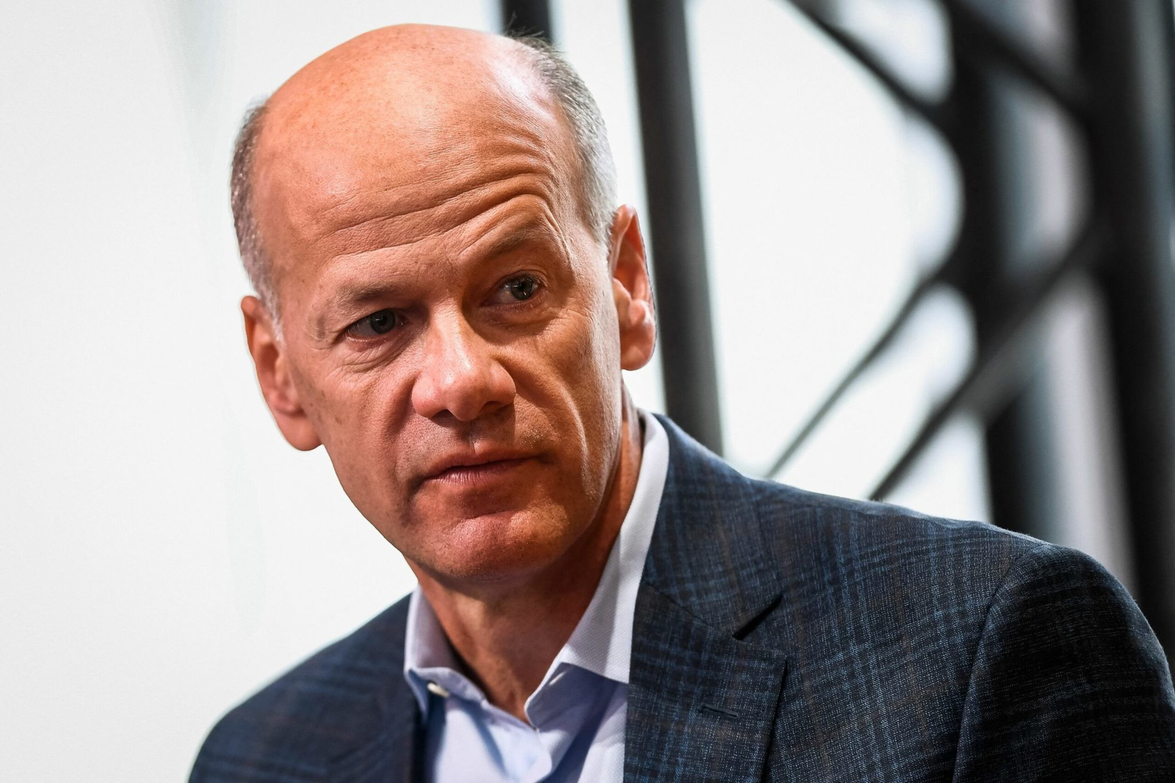A middle-aged man with a bald head is wearing a dark checkered blazer over a white collared shirt, looking to his left with a serious expression.