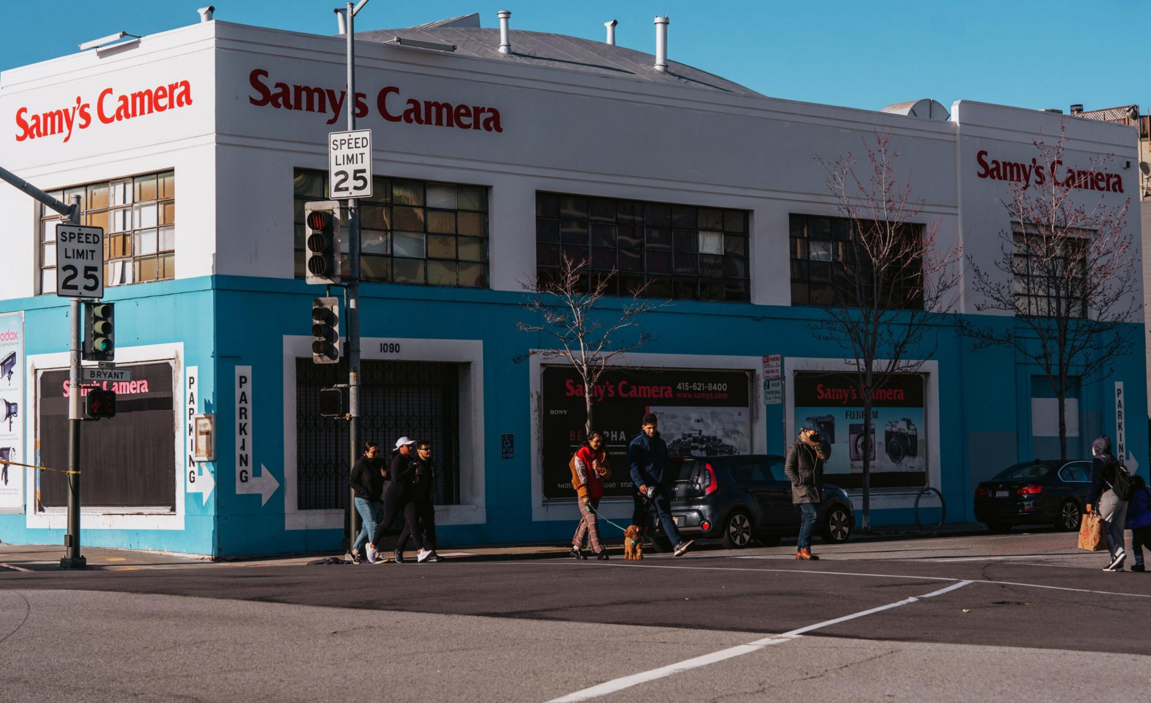 A blue-and-white building with "Samy's Camera" signs stands at a street corner, with several pedestrians and cars nearby under clear skies.