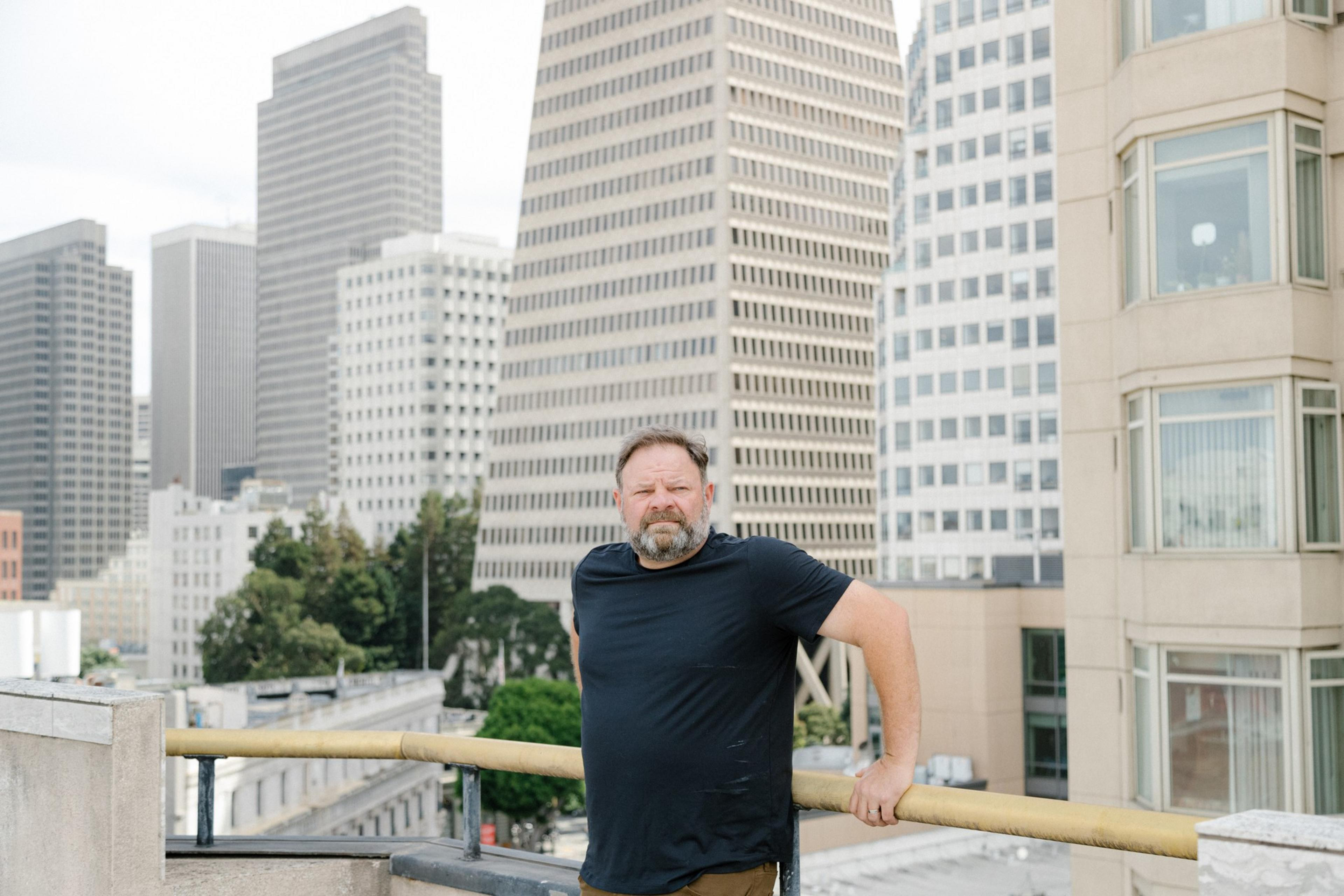 A man with a beard and black shirt leans against a rooftop railing with tall city buildings and trees in the background.