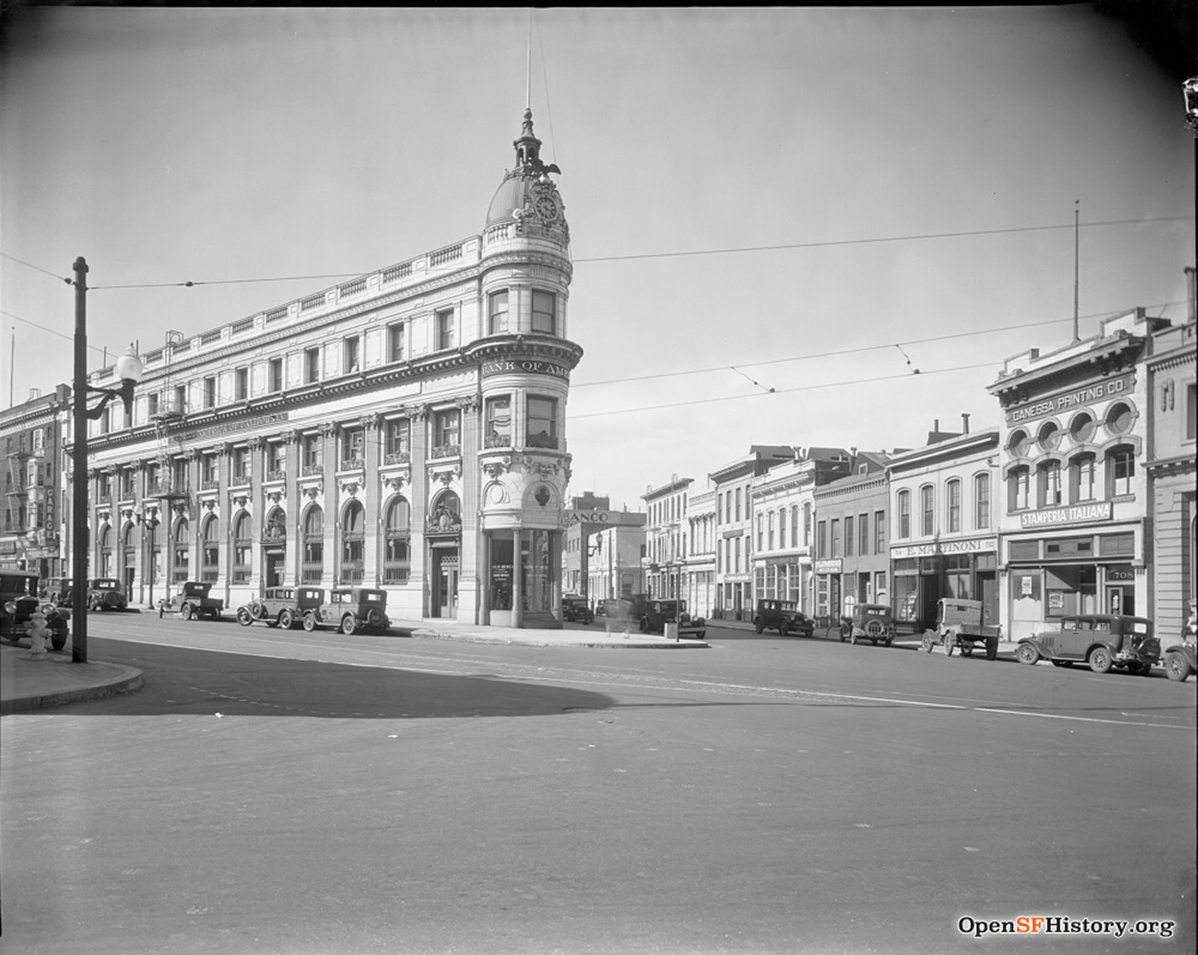 A historic street scene features a tall Bank of America building with a clock tower, vintage cars parked along the street, and smaller storefronts including a printing company.