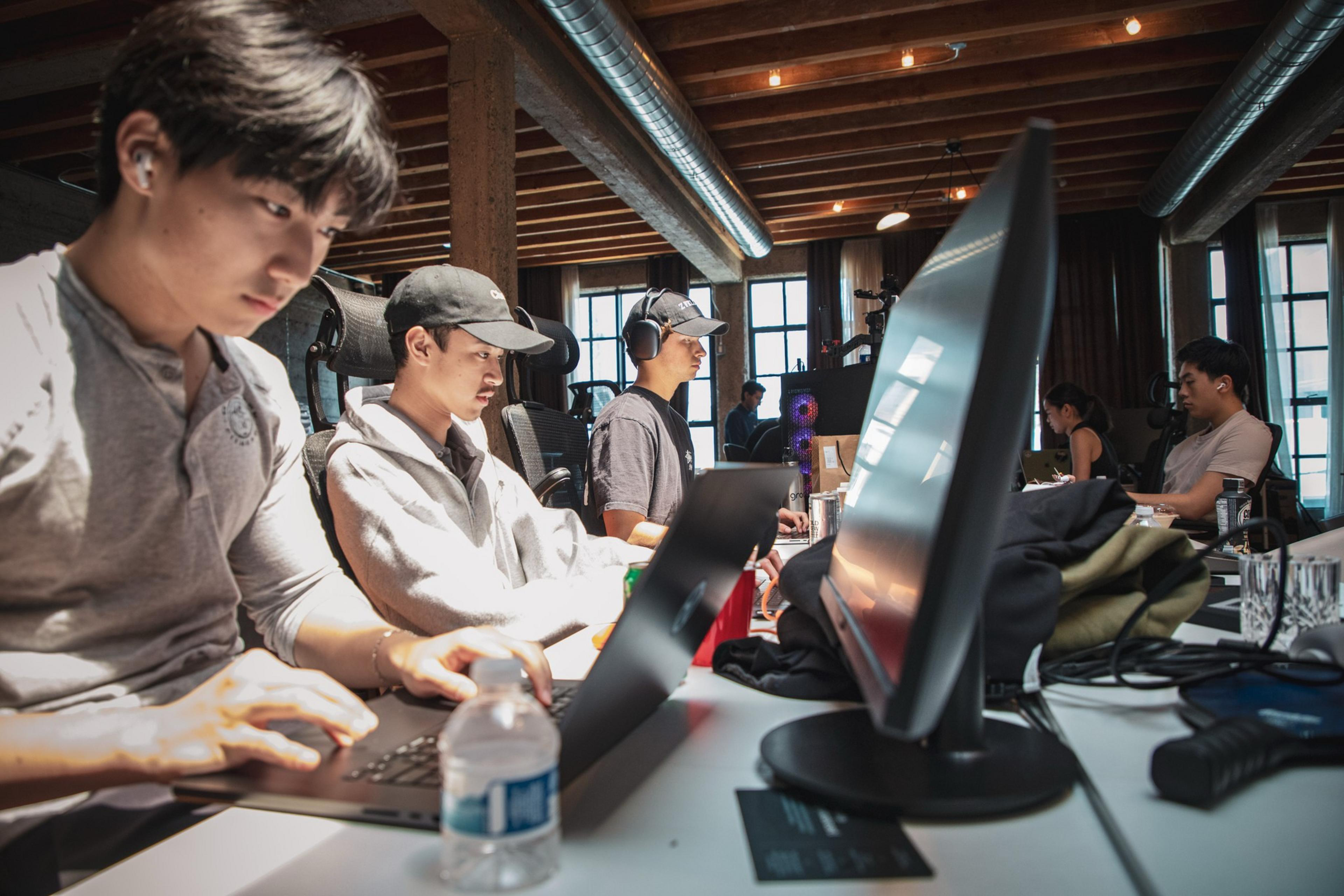 A group of people are working at computers in a modern, industrial-style office with wooden beams and exposed ducts. They appear focused and engaged.