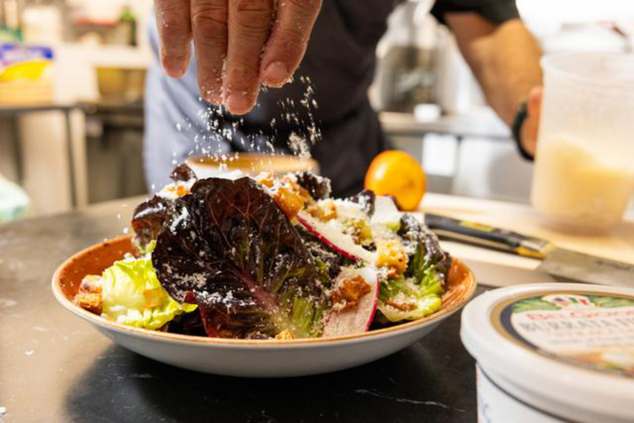 A hand sprinkles grated cheese over a fresh salad with leafy greens, radish slices, and croutons on a plate in a kitchen setting.