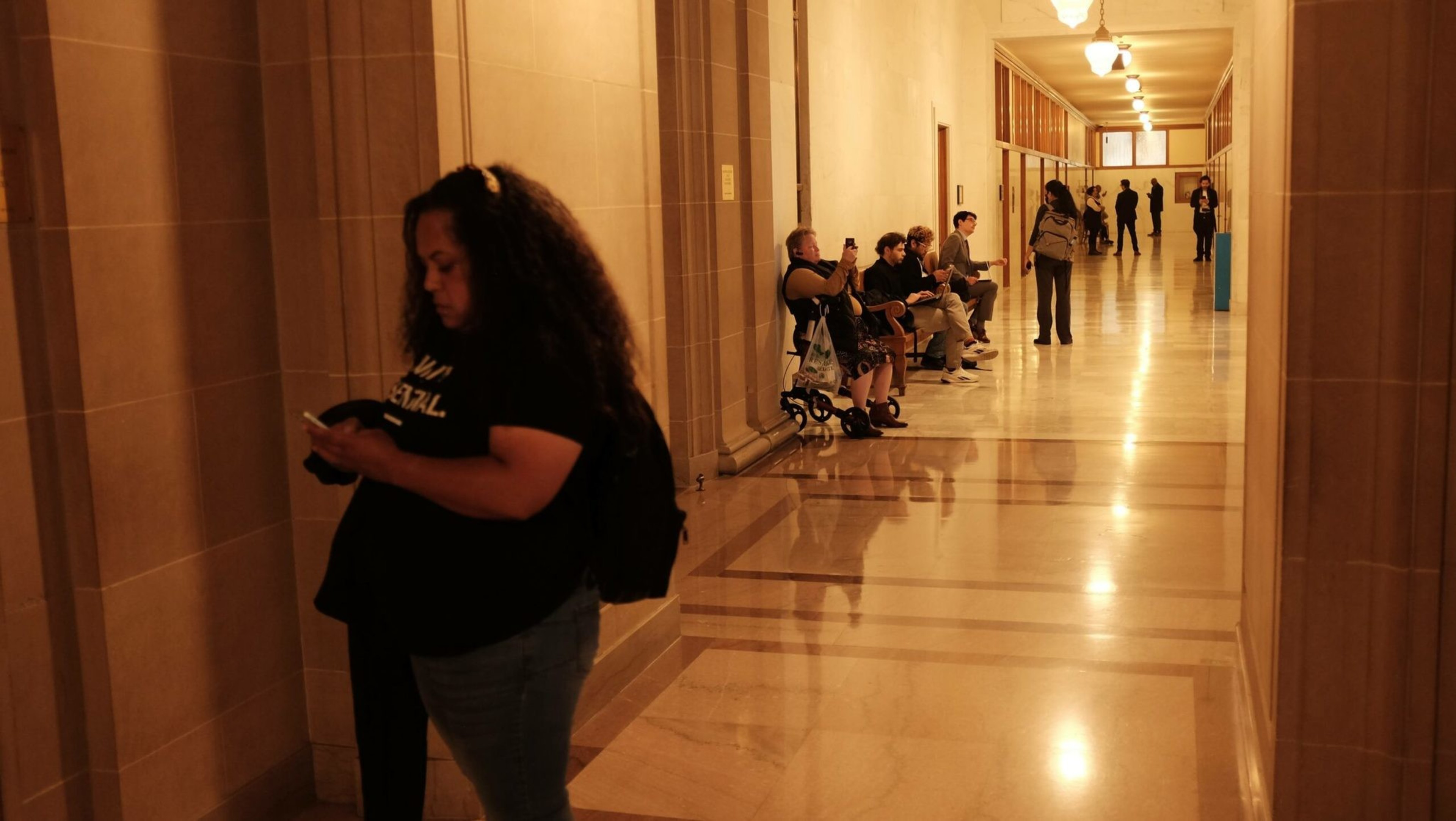A woman with curly hair stands looking at her phone in a dimly lit hallway where several people sit or stand along the walls.