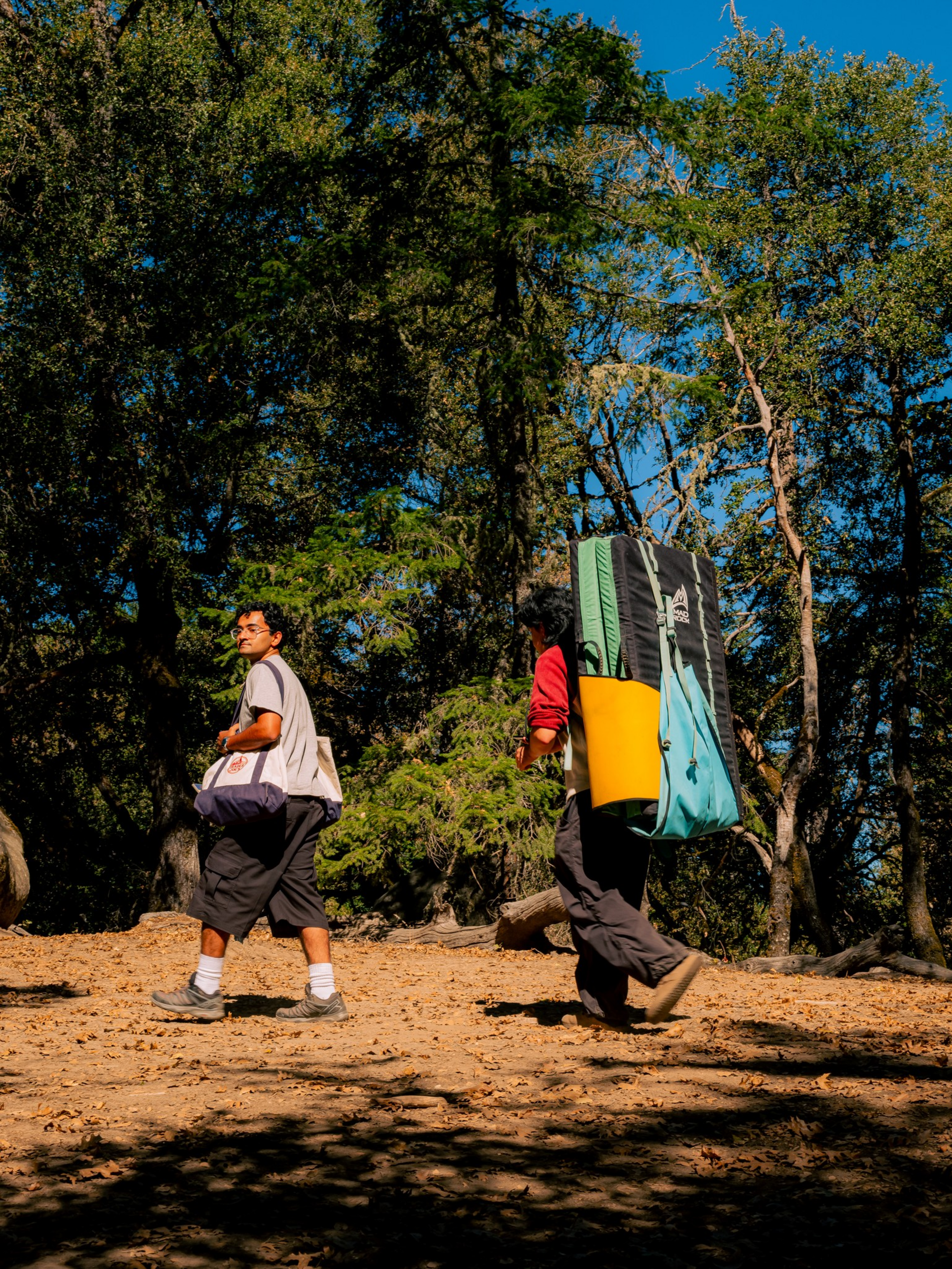 Two hikers walk on a dirt path through a sunlit forest, one carrying a large blue and yellow backpack and the other holding a smaller bag.