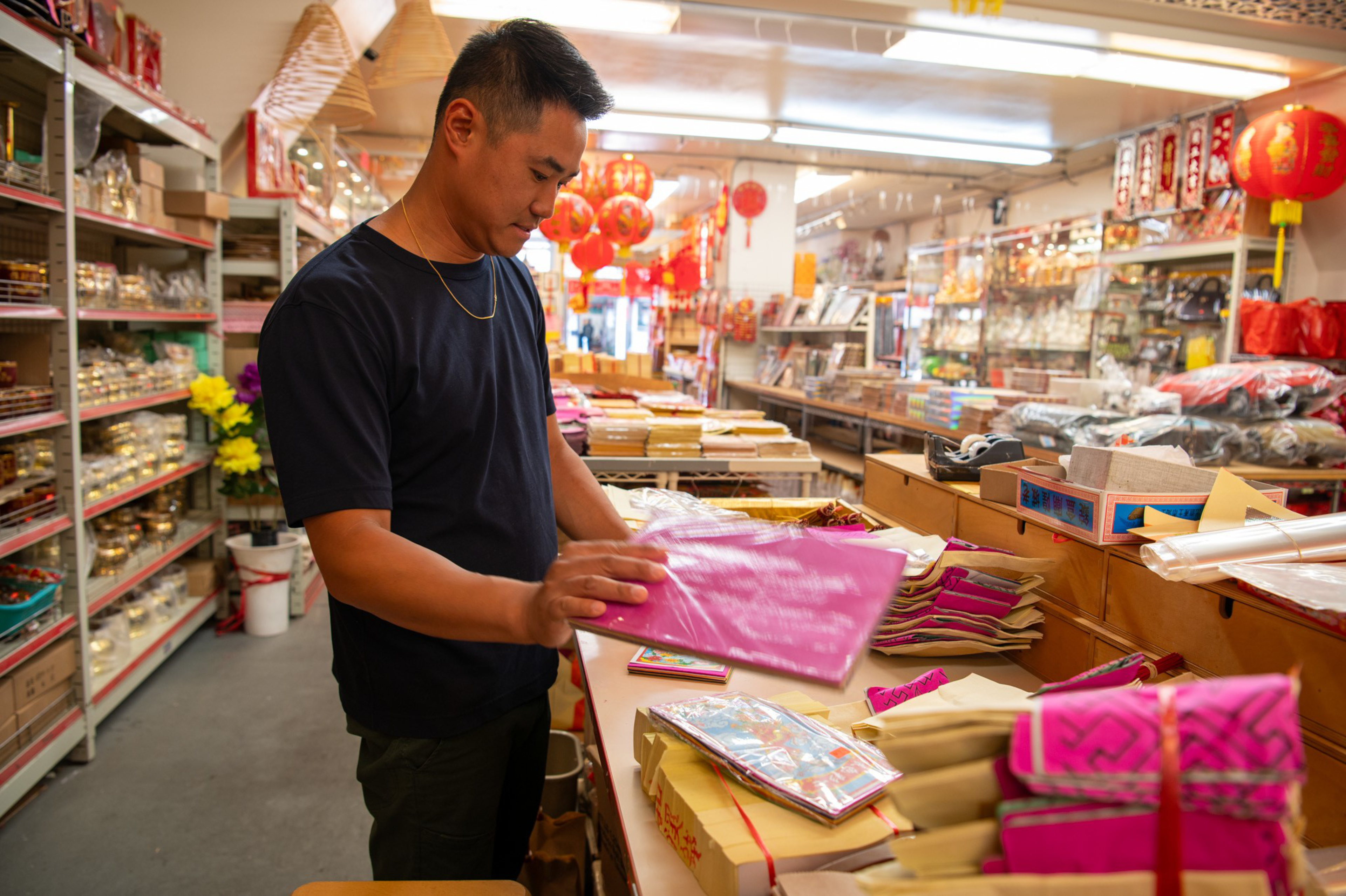 A man in a black shirt browses pink paper sheets inside a brightly lit store decorated with red lanterns and filled with various packaged goods.