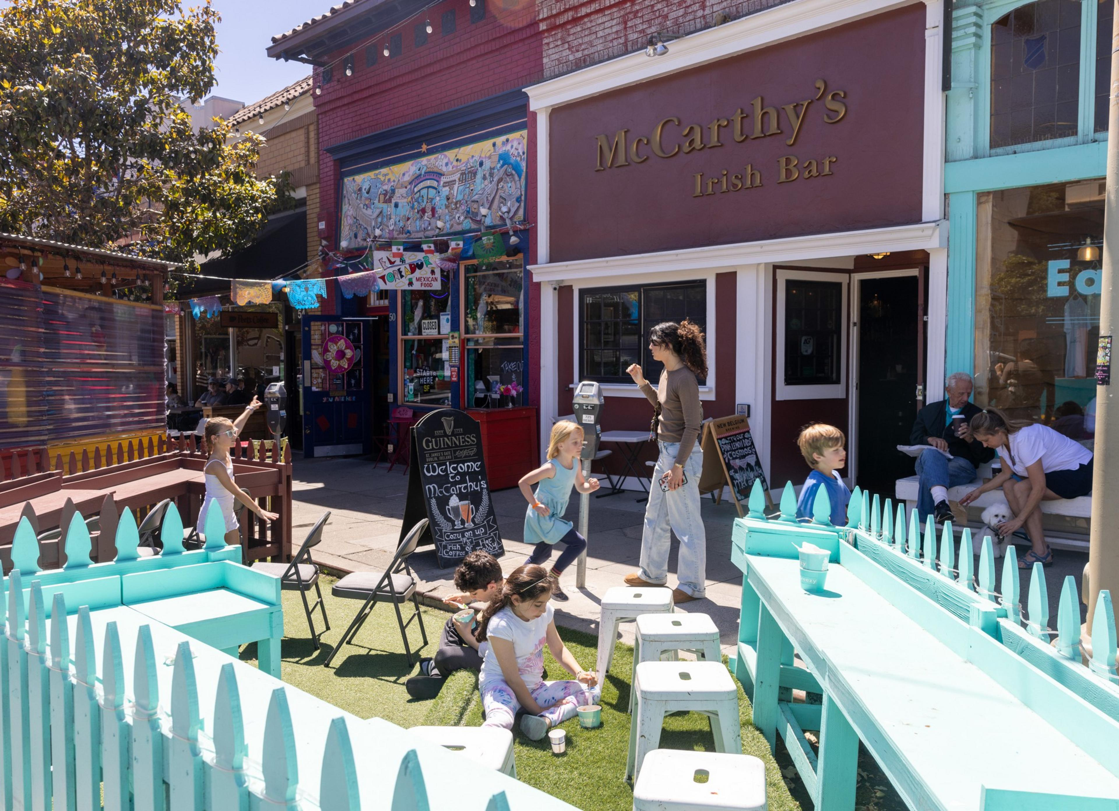 Children and adults socialize outside McCarthy's Irish Bar. Bright decor, a blue fence with benches, and colorful storefronts add vibrant energy to the street scene.