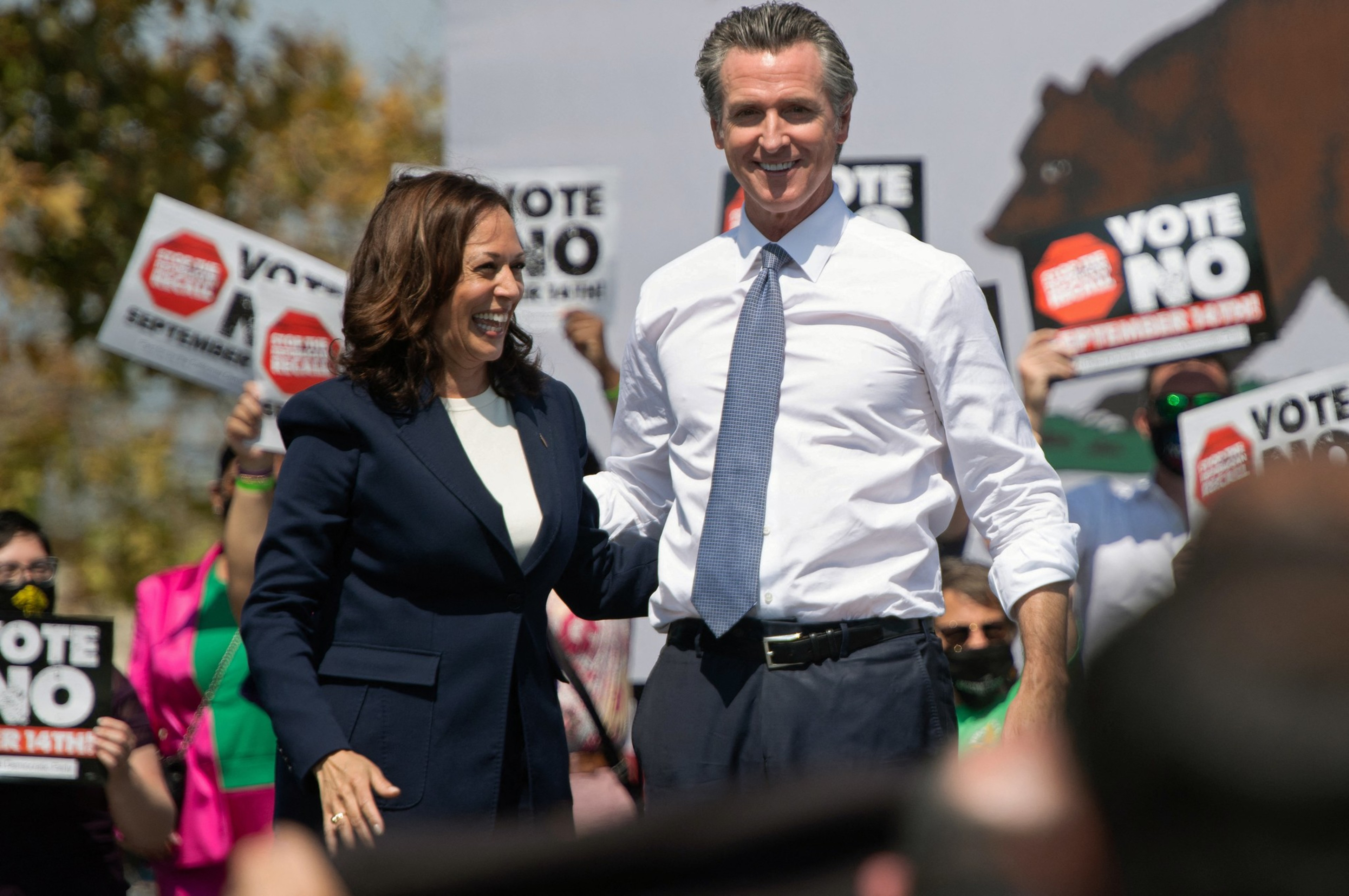 A woman in a dark suit and a man in a white shirt with rolled-up sleeves and blue tie smile as they stand with their arms around each other, while people behind hold "VOTE NO" signs.