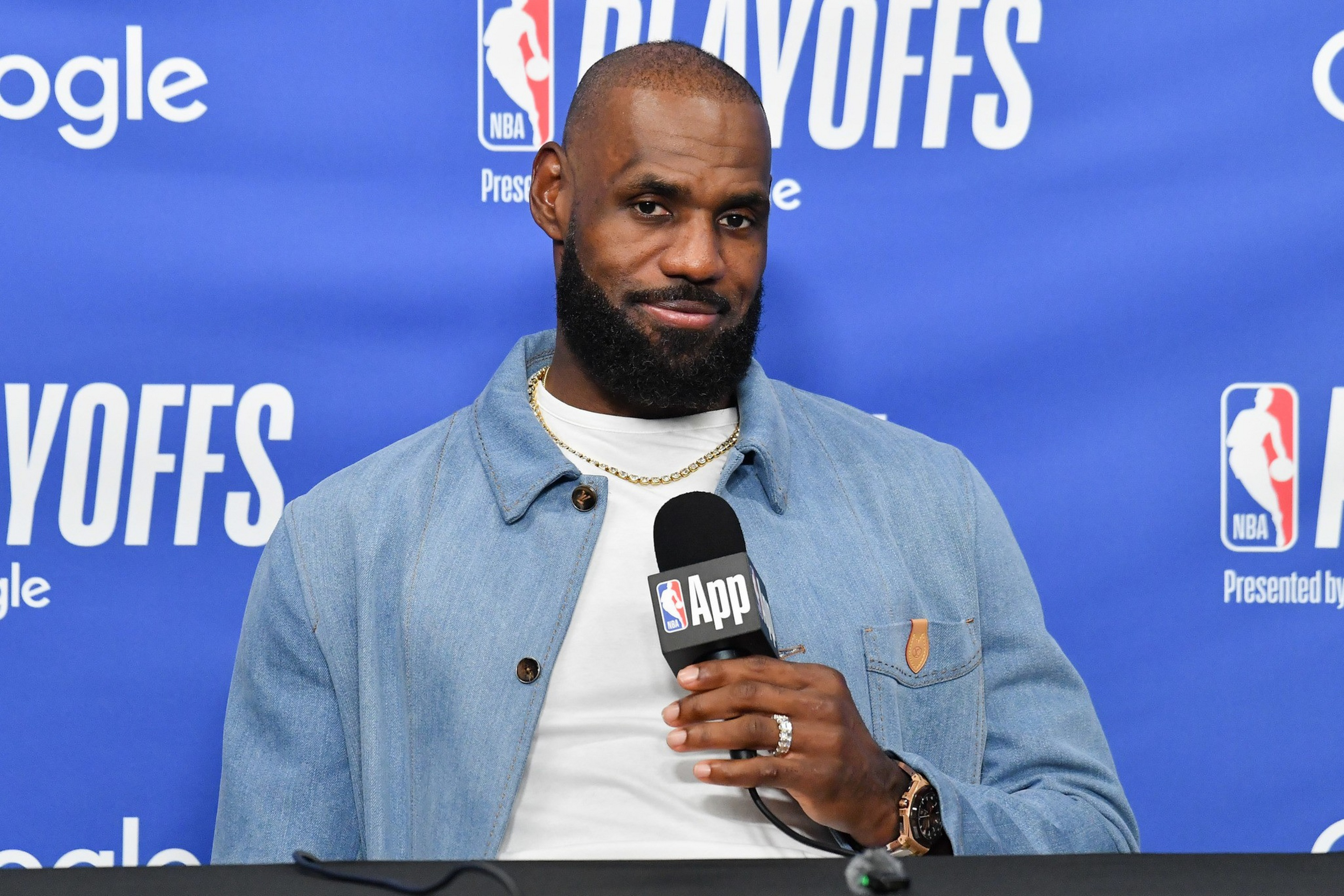A man with a beard and gold chain holds an NBA App microphone, sitting at a table against a blue NBA Playoffs backdrop.
