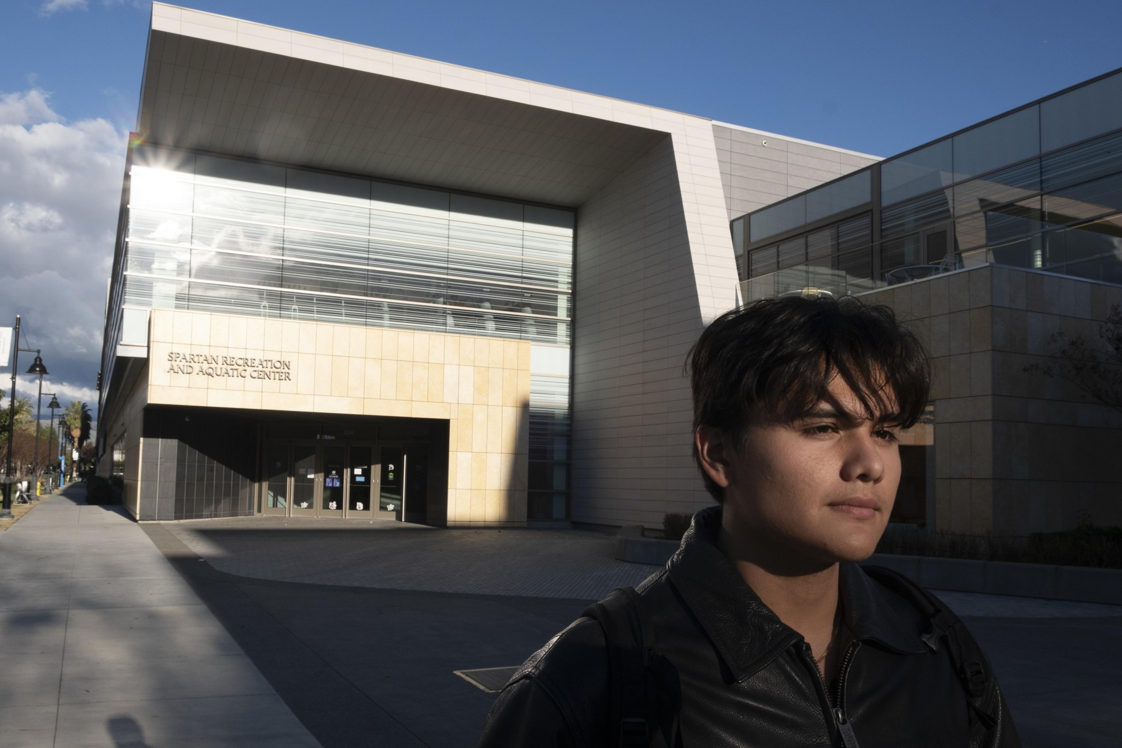 A young person in a black jacket stands in front of the modern Spartan Recreation and Aquatic Center building, with sunlight reflecting off its glass facade.
