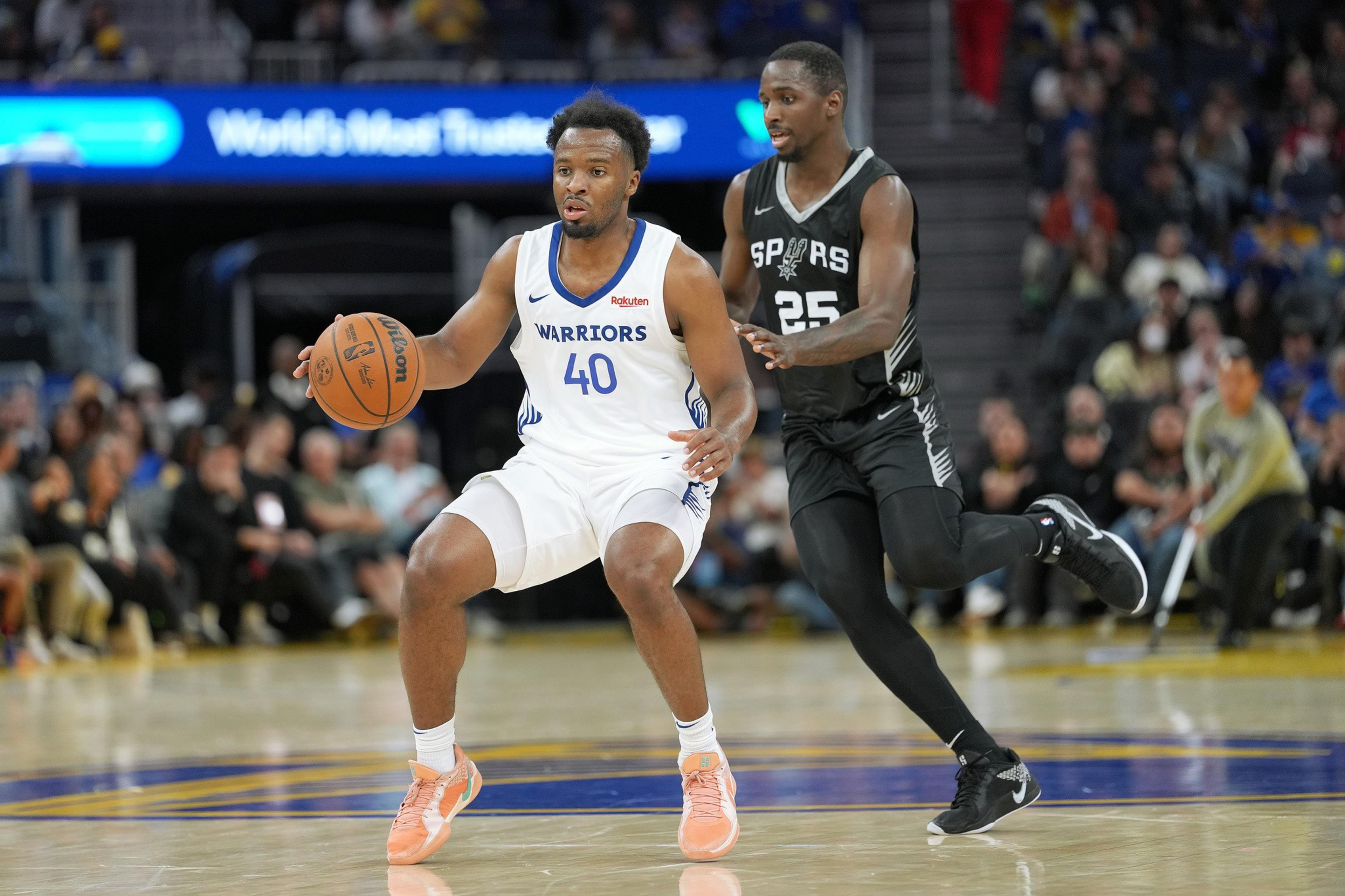 A basketball player in a Warriors jersey dribbles the ball while being guarded by a Spurs player. The game is taking place on a brightly lit court.