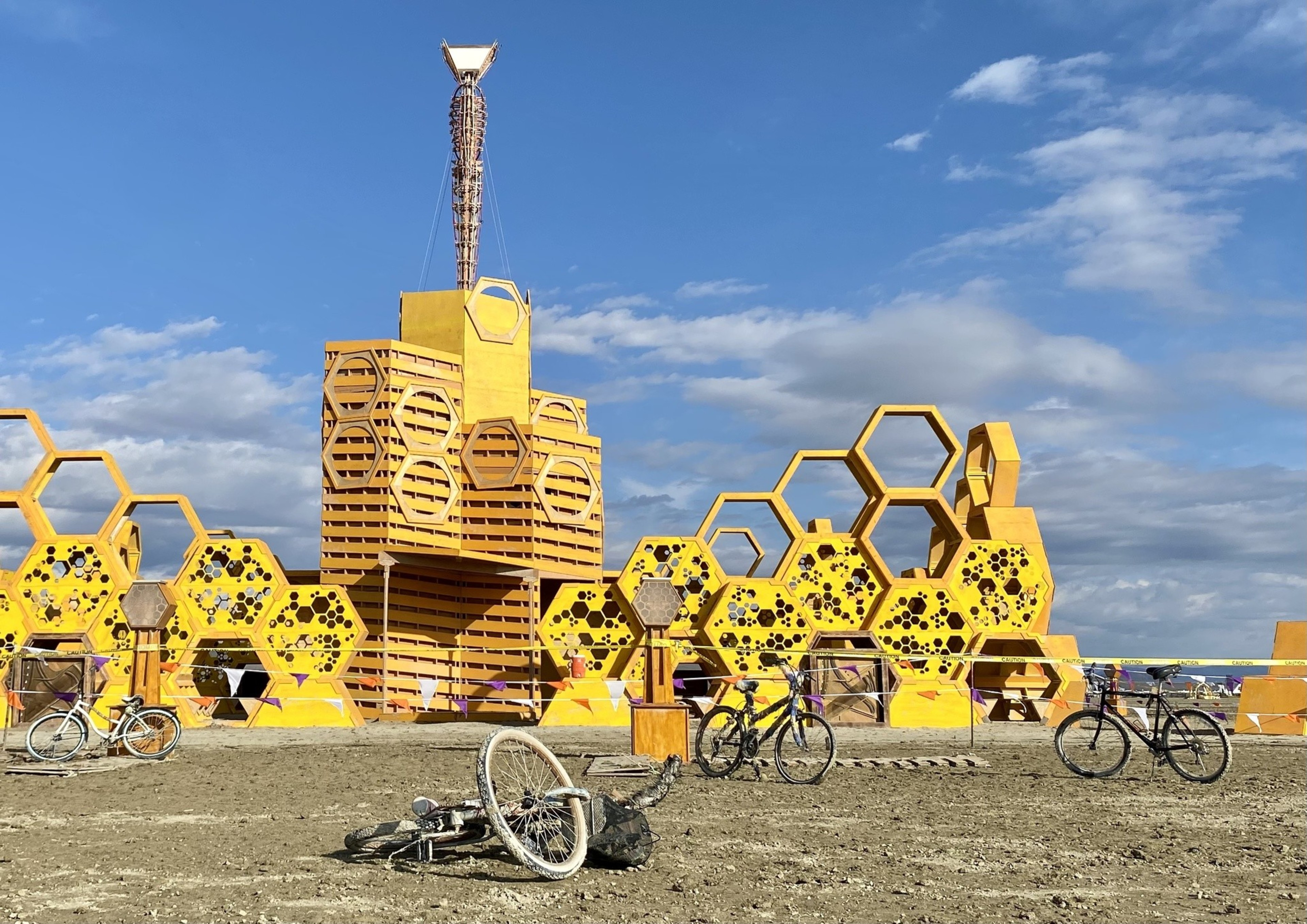 A yellow hexagon-themed structure with a tall central column, surrounded by bicycles on a dry, dirt ground under a bright, cloudy sky.