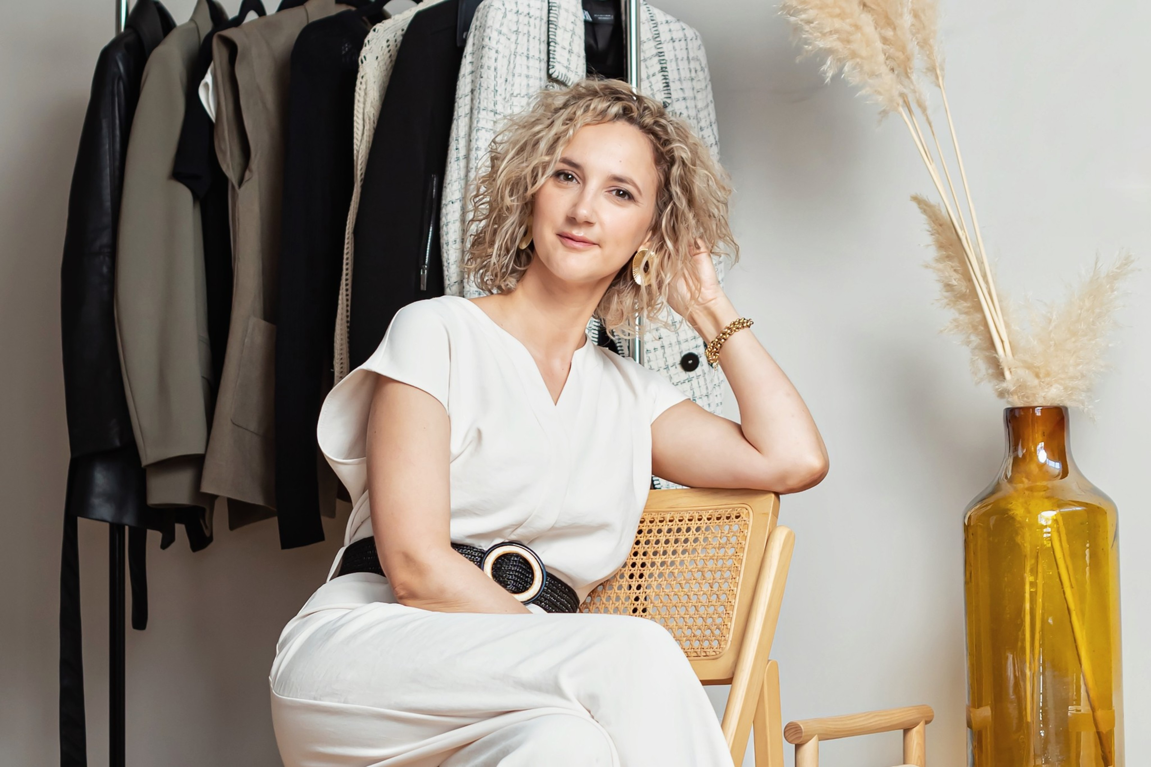 San Francisco stylist Lili Henry sitting on a wicker chair with a rack of clothes behind her and a large vase with pampas grass