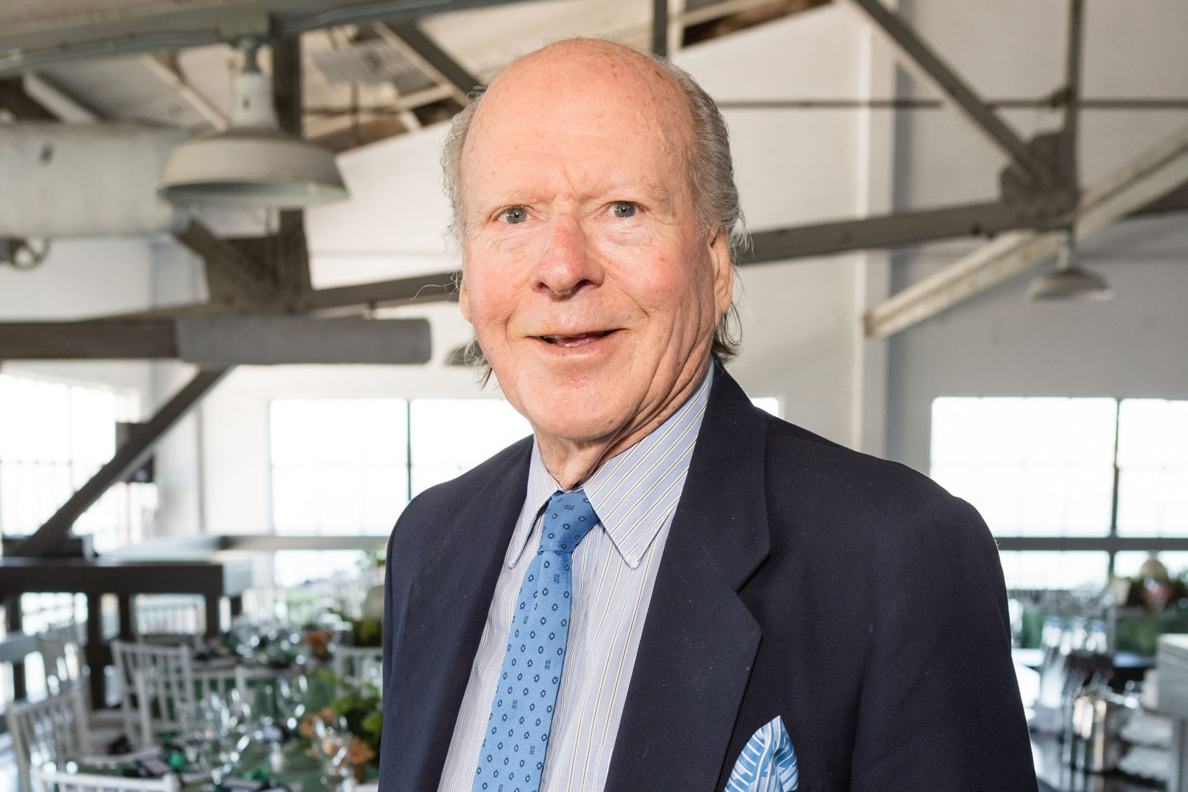 An elderly man wearing a blue suit, striped shirt, and blue tie smiles in a sunlit room with exposed beams and set dining tables.