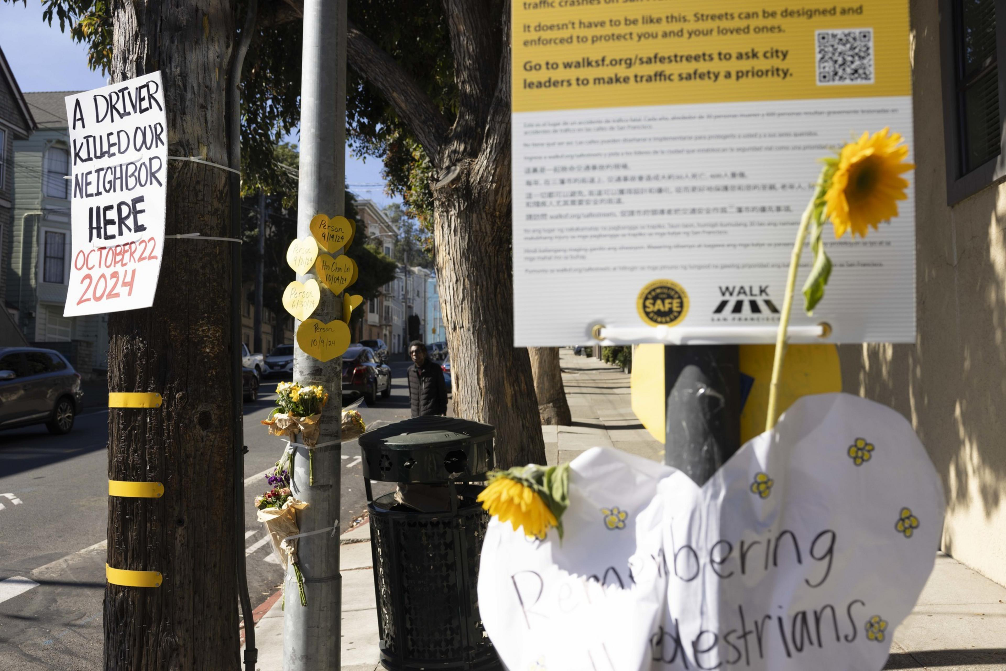A memorial on a street features flowers and signs about pedestrian safety, including one with the date October 22, 2024, noting a tragic incident.