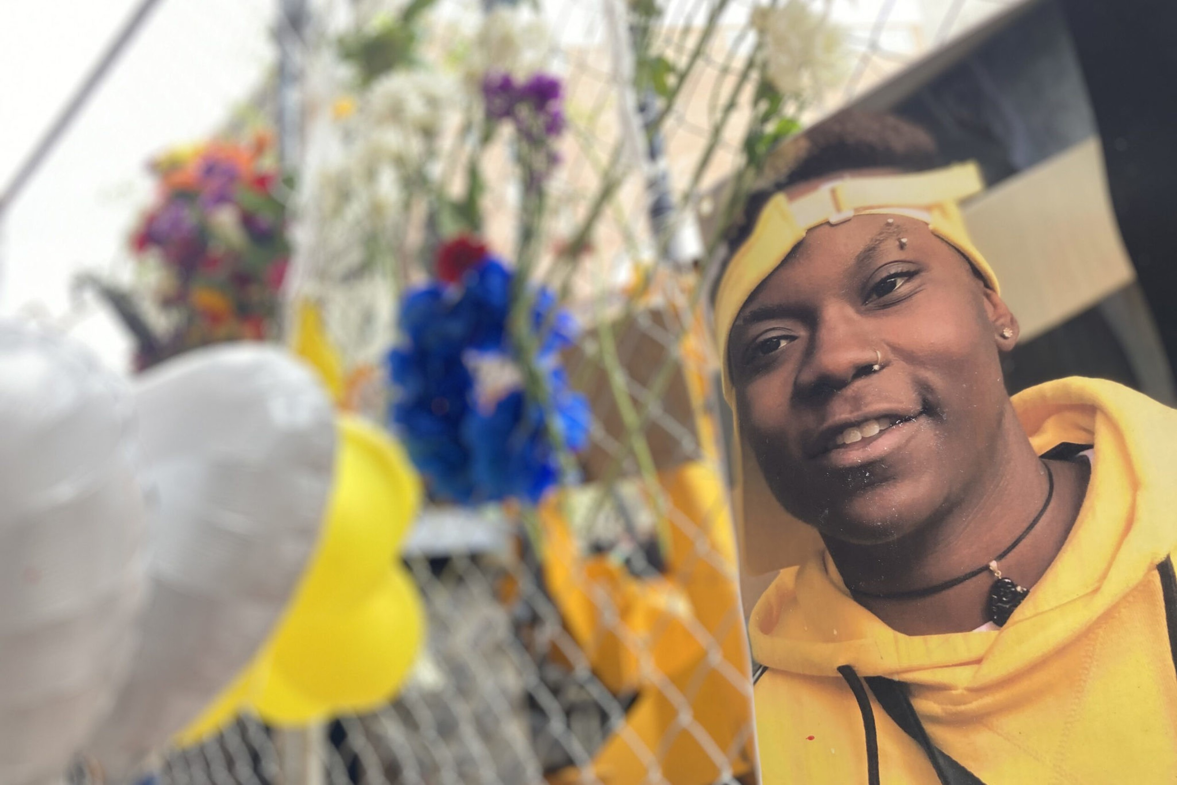 A smiling person wearing a yellow headband and hoodie is pictured on a memorial fence decorated with colorful flowers and balloons.