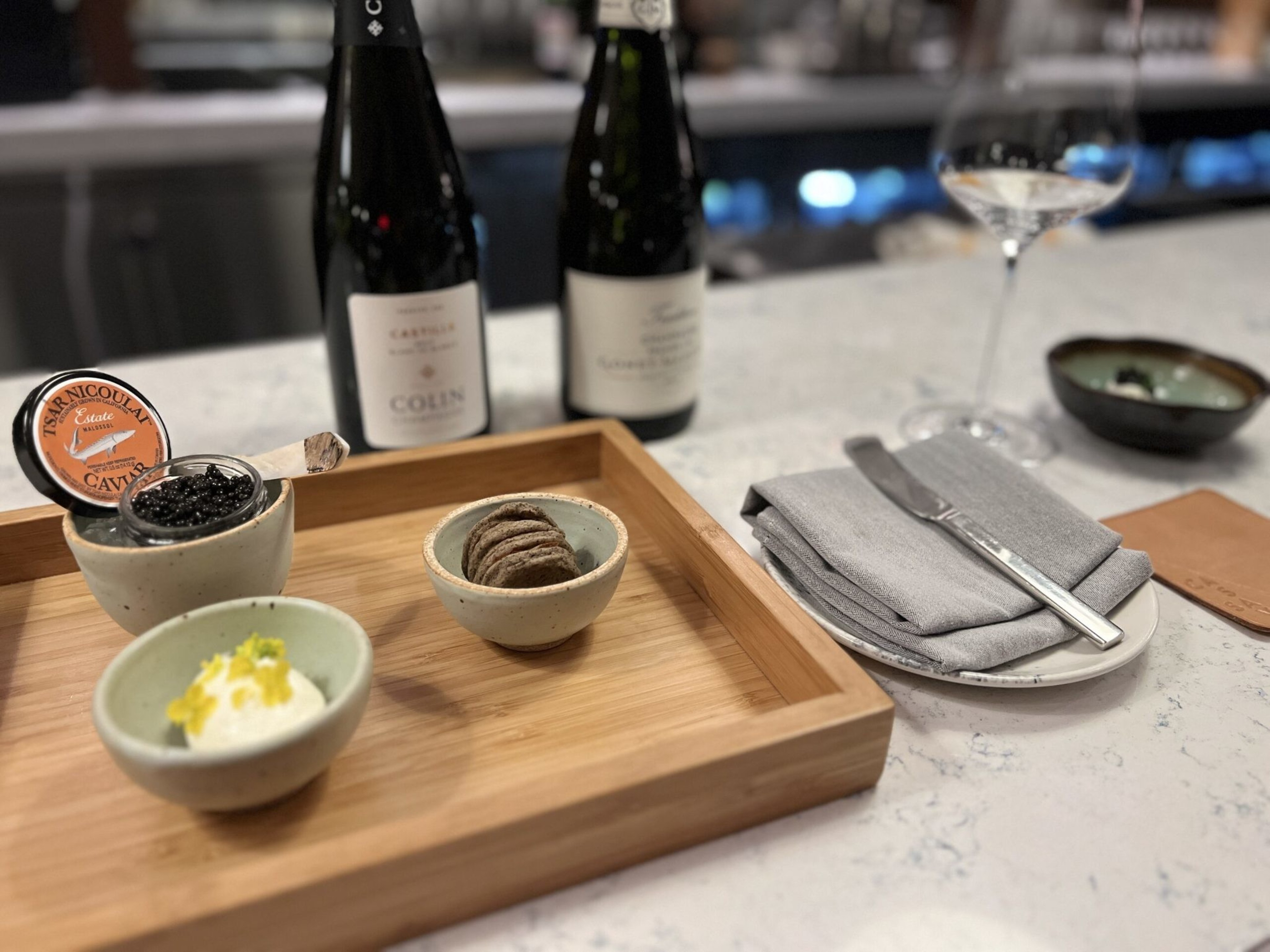 A wooden tray holds small bowls with black caviar, crackers, and a creamy spread, next to two wine bottles, a wine glass, and a plate with a folded gray napkin and knife.