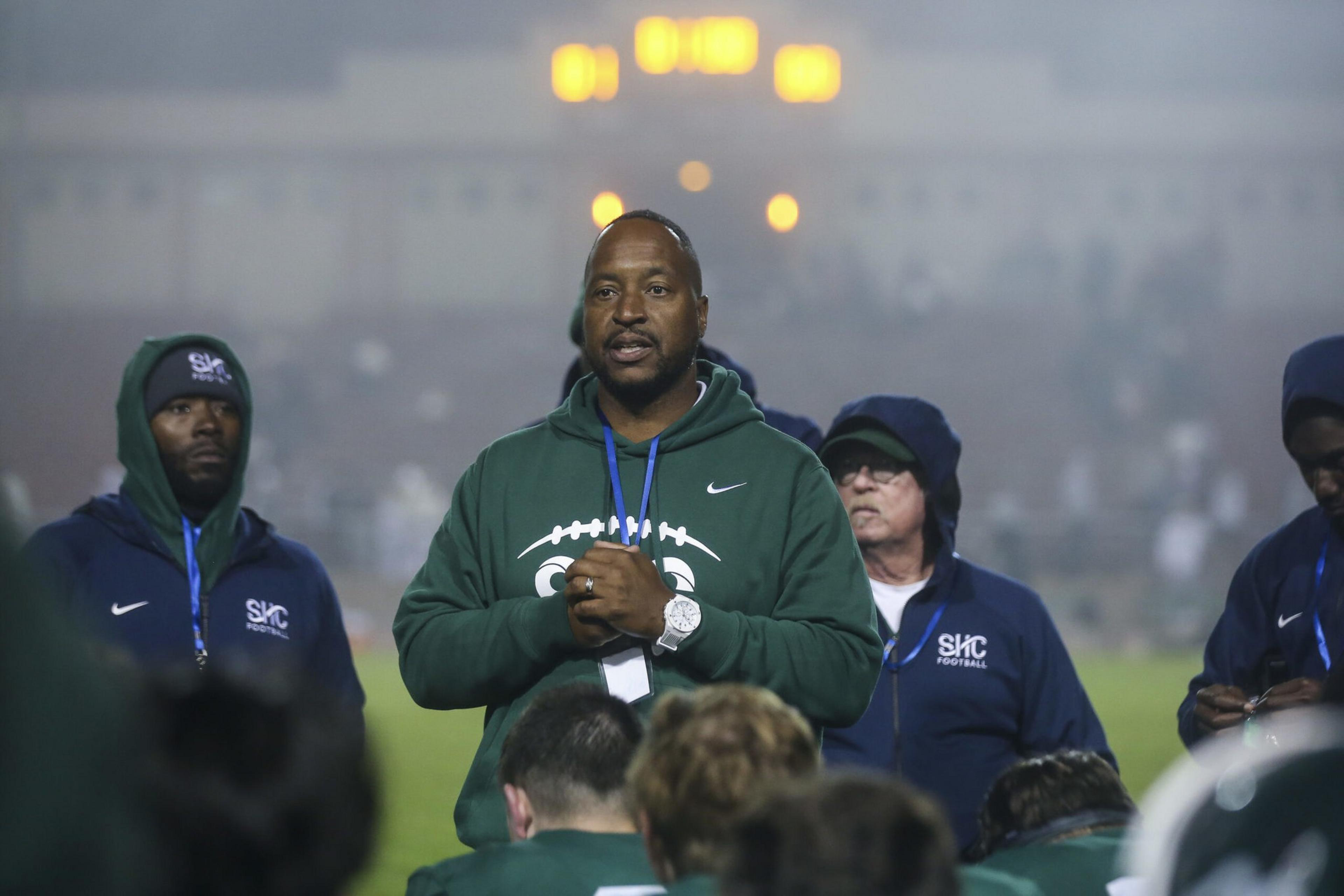 A coach wearing a green hoodie stands speaking to players, flanked by assistants in navy blue SKC Football jackets under stadium lights.