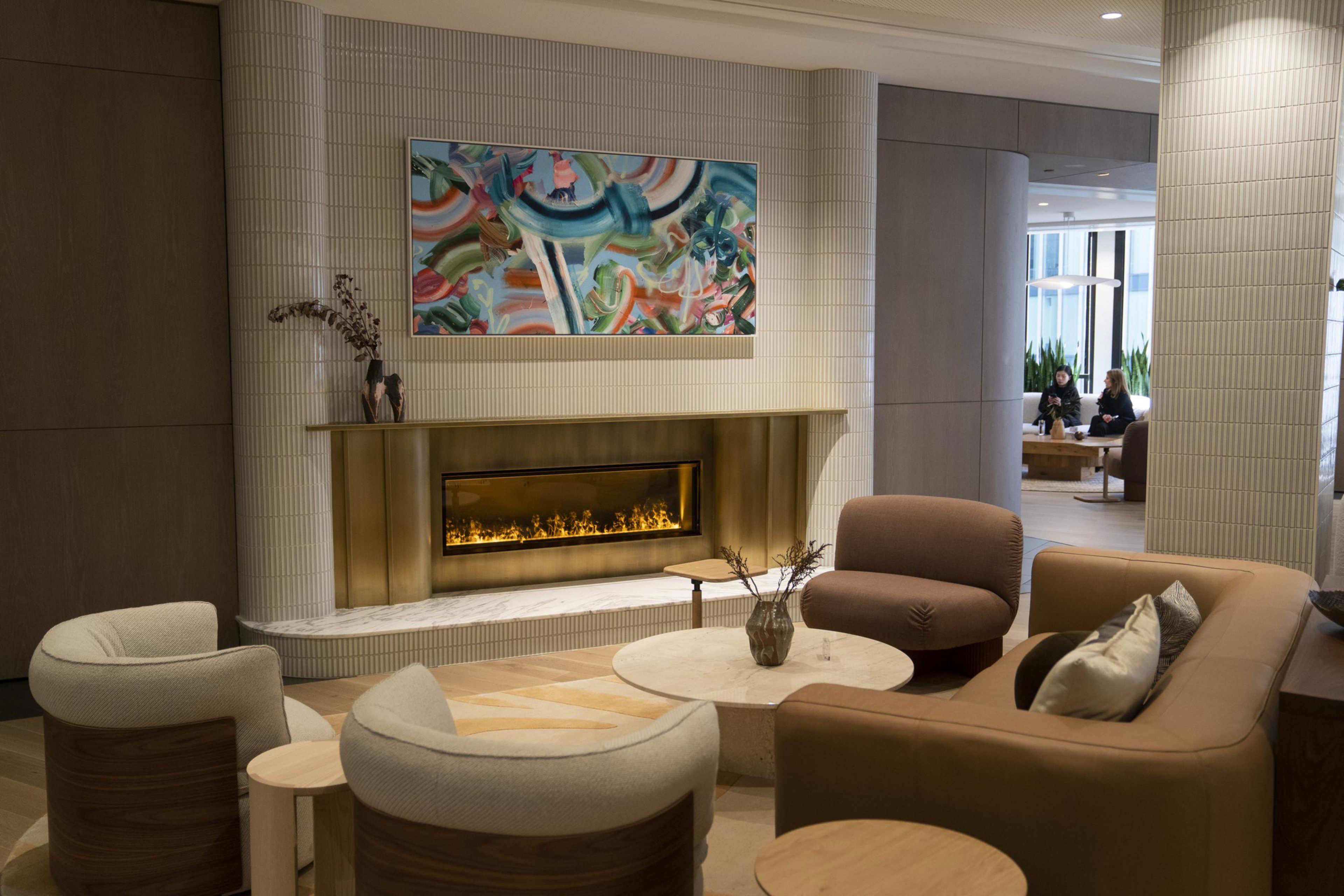 A cozy lounge features a modern fireplace, abstract art above, plush seating, and a round table with a decorative vase. Two people are seated in the background.