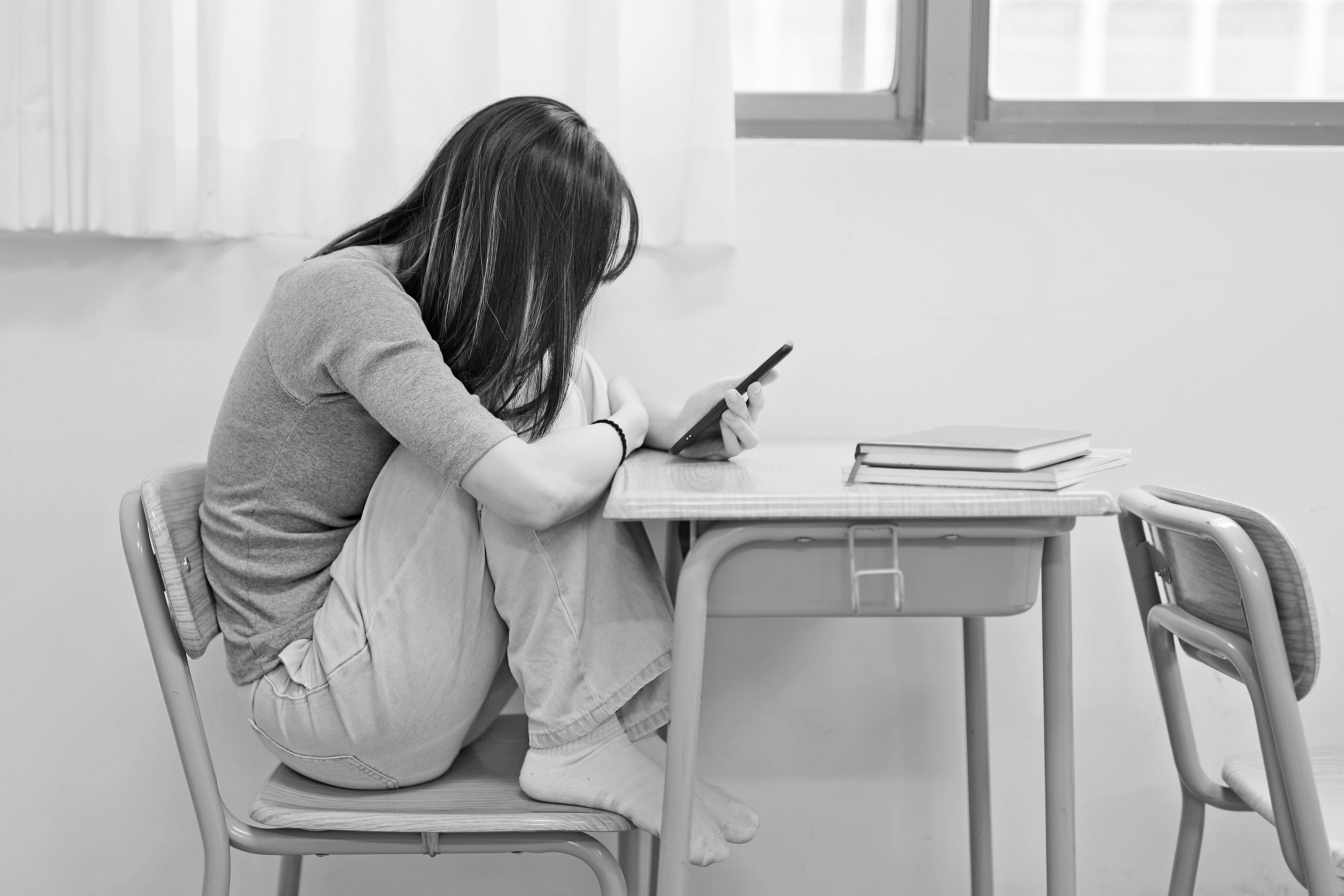 A person sits curled up on a chair at a school desk, looking at a smartphone, with two books placed on the desk, in a seemingly contemplative or stressed posture.