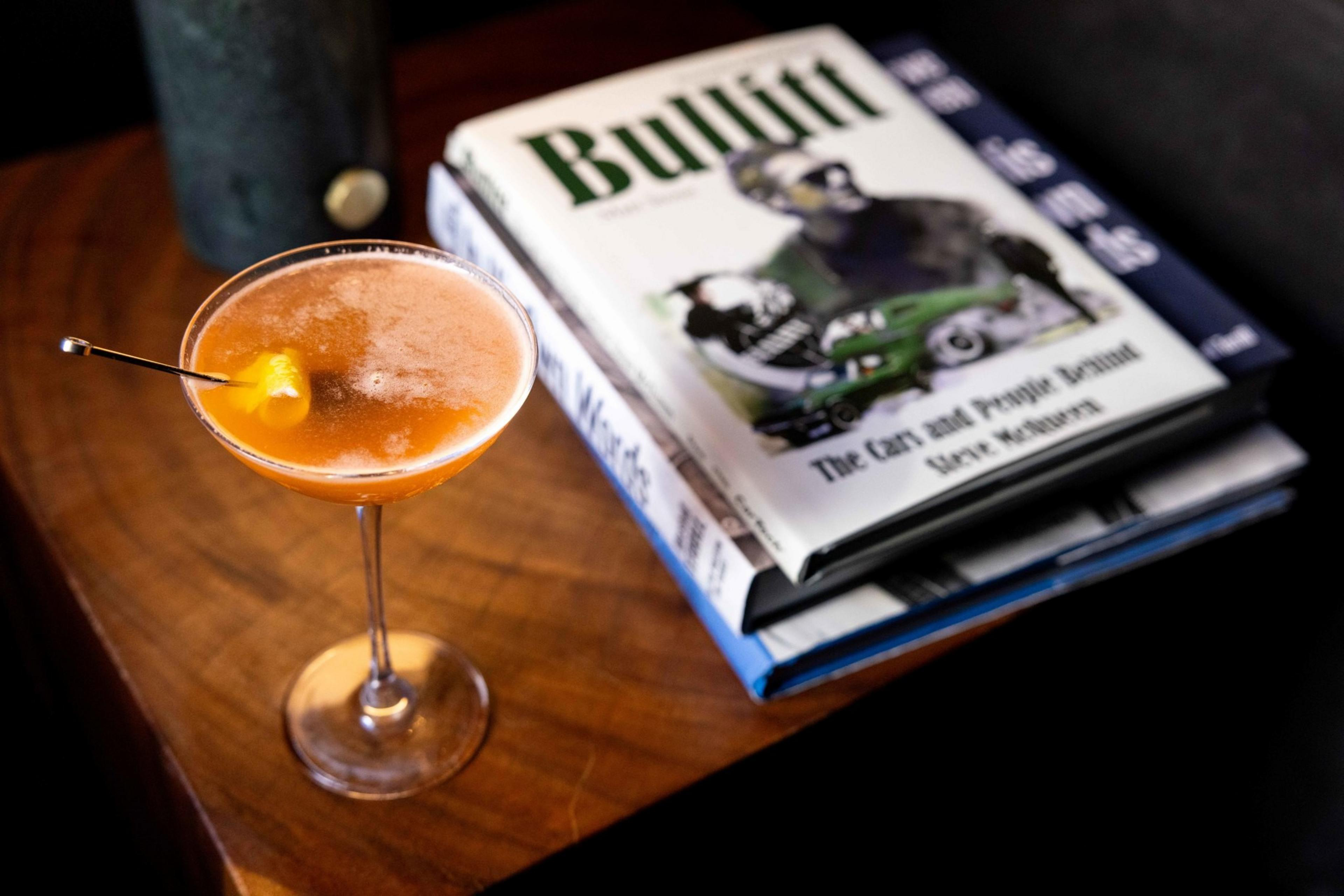A cocktail with an orange hue and cherry garnish sits on a wooden table. Nearby, there's a stack of books, with one titled &quot;Bullitt&quot; on top.