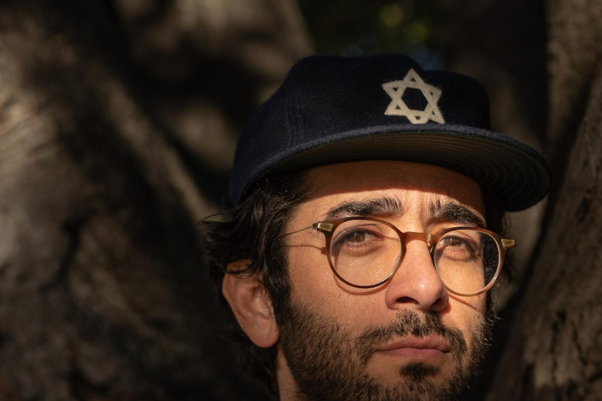 A man with glasses and a beard stands in dappled sunlight. He wears a denim shirt, beige cardigan, and a dark cap with a Star of David. Dark tree trunks frame him.