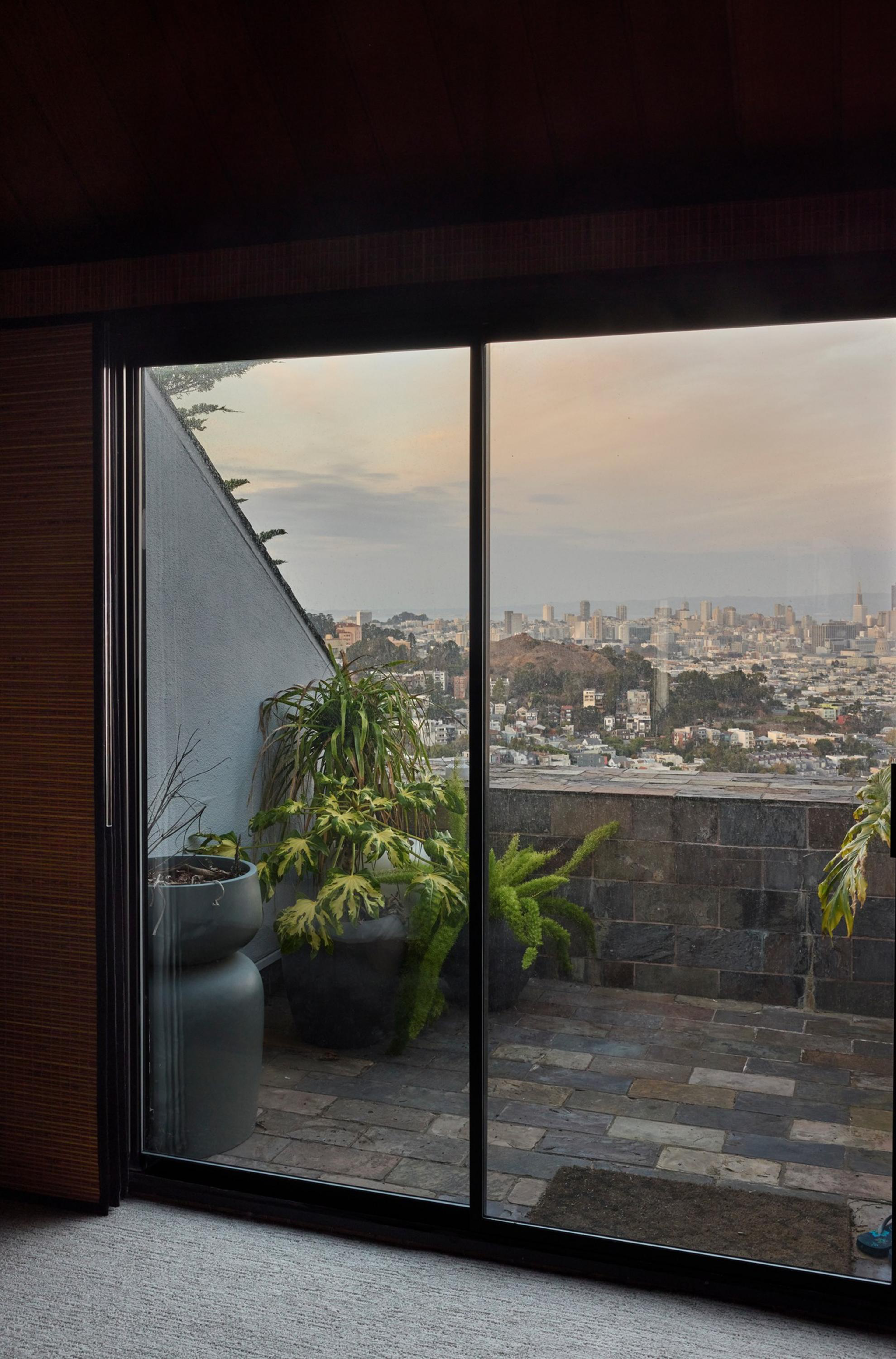 A balcony with potted plants overlooks a cityscape under a soft, cloudy sky seen through large sliding glass doors.