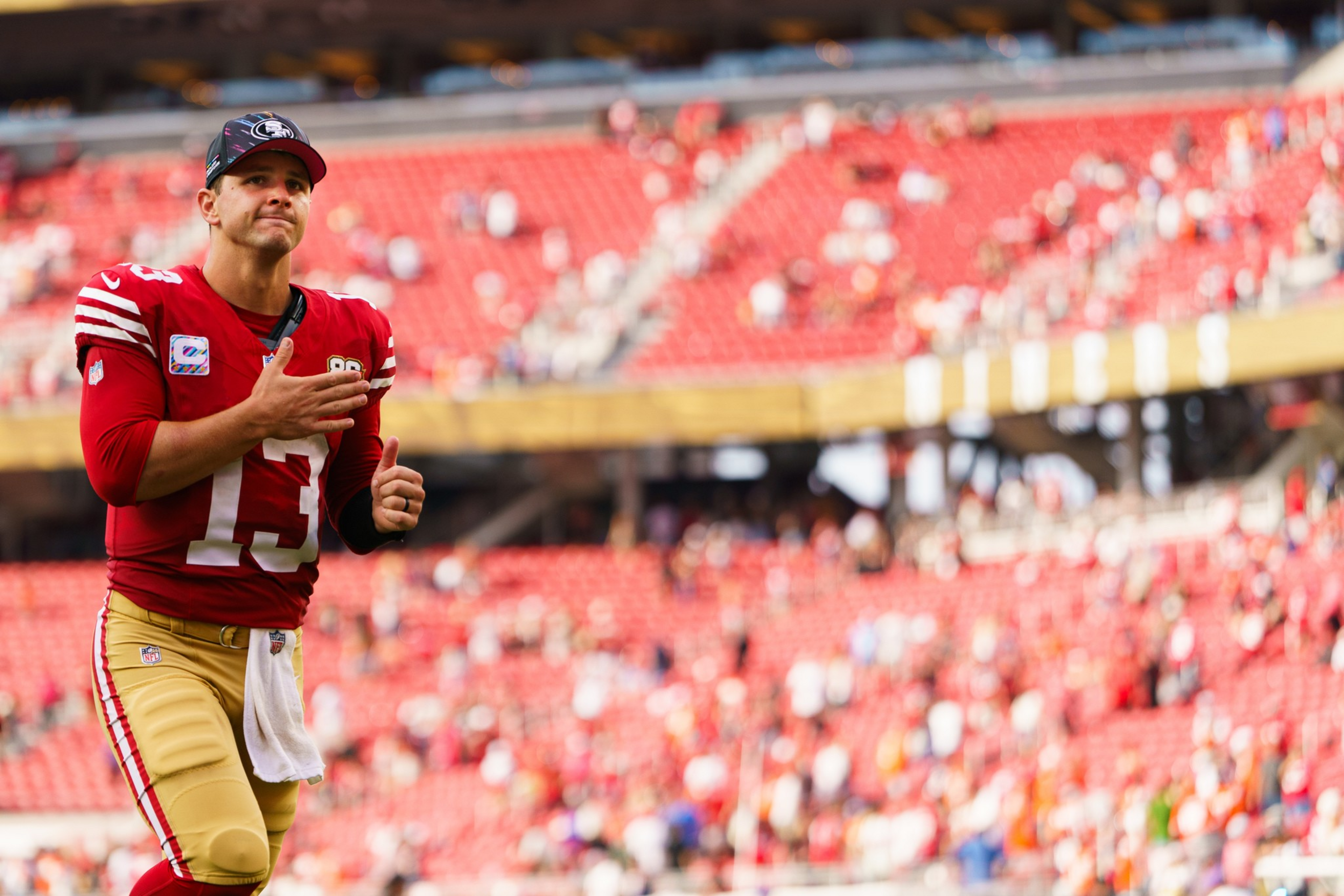 A football player in a red and gold uniform with number 13 is walking while clapping, with empty stadium seats and a scattered crowd in the background.