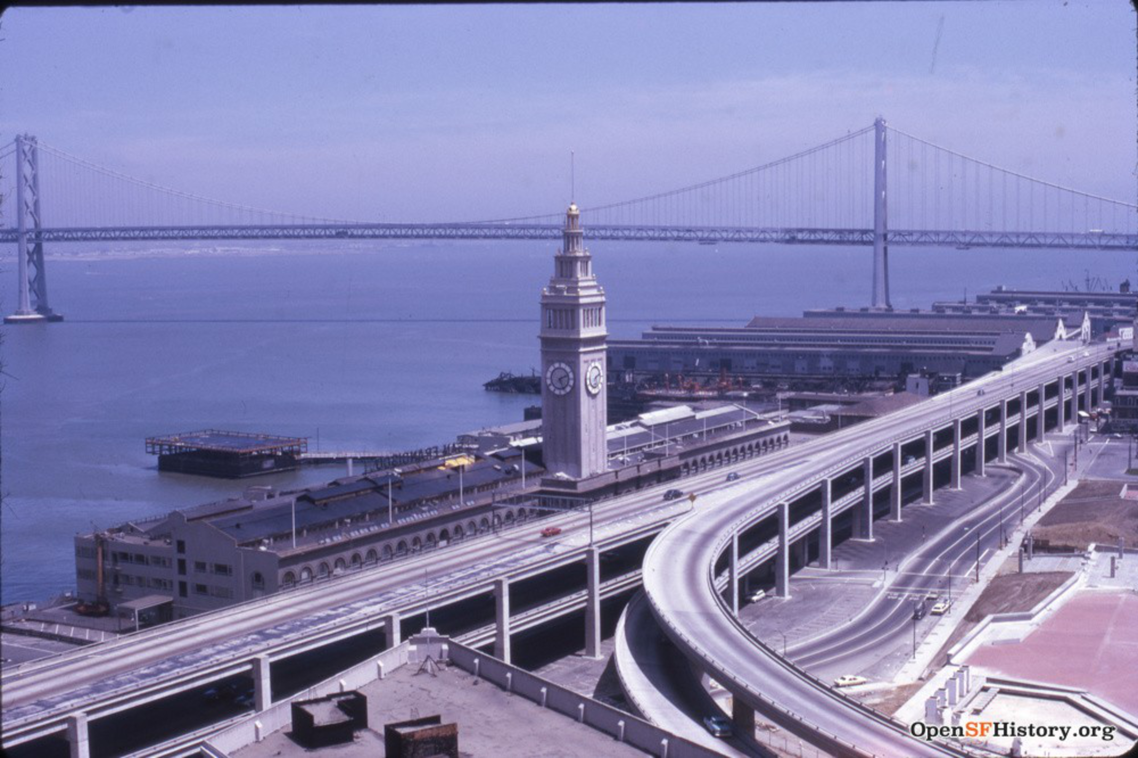 A clock tower stands near a waterfront with elevated highways curving alongside, and a large suspension bridge spans across the water in the background.