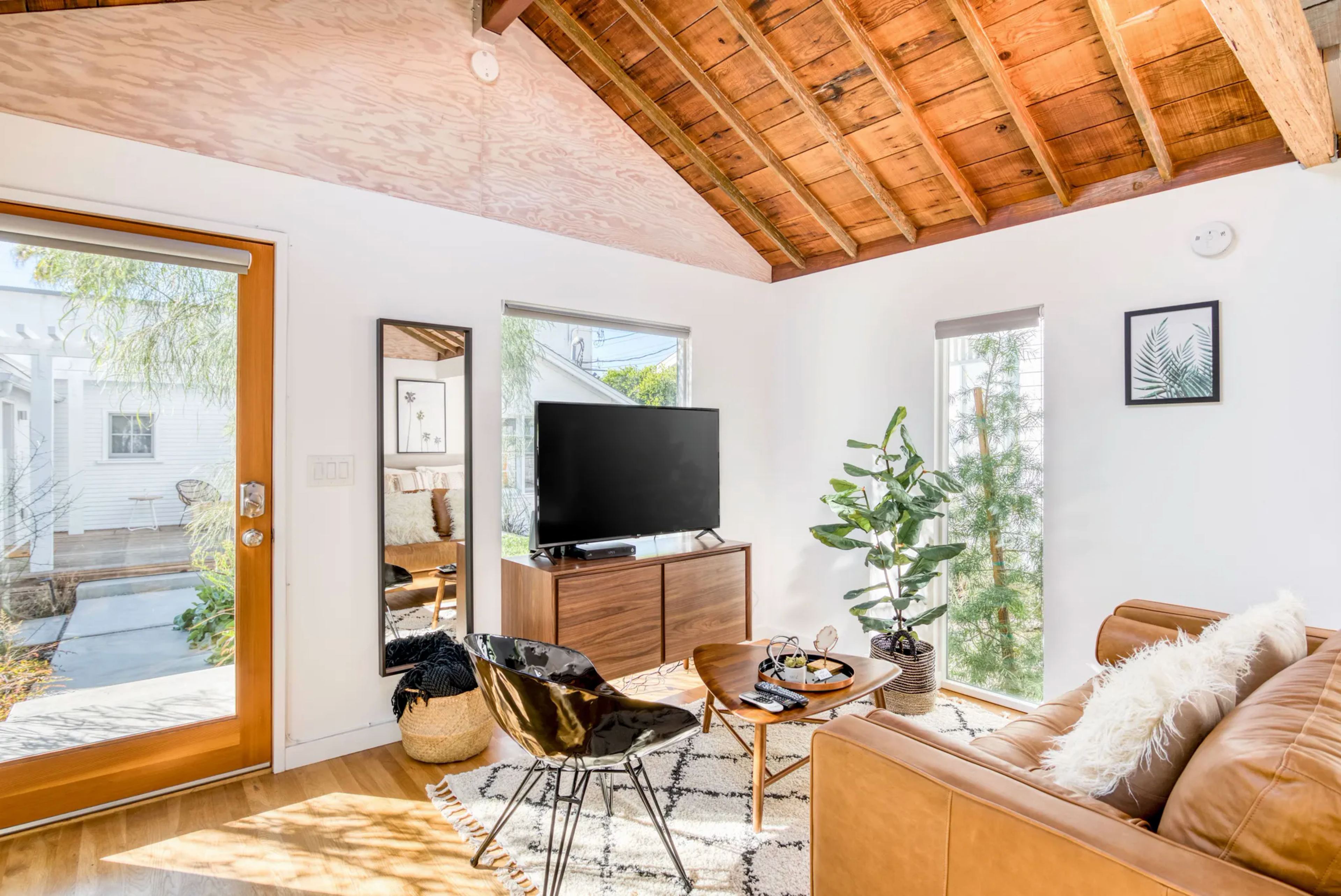 A picture of a living room shows a mid century modern chair, a TV stand with a TV on it, plants and windows with a wood ceiling.