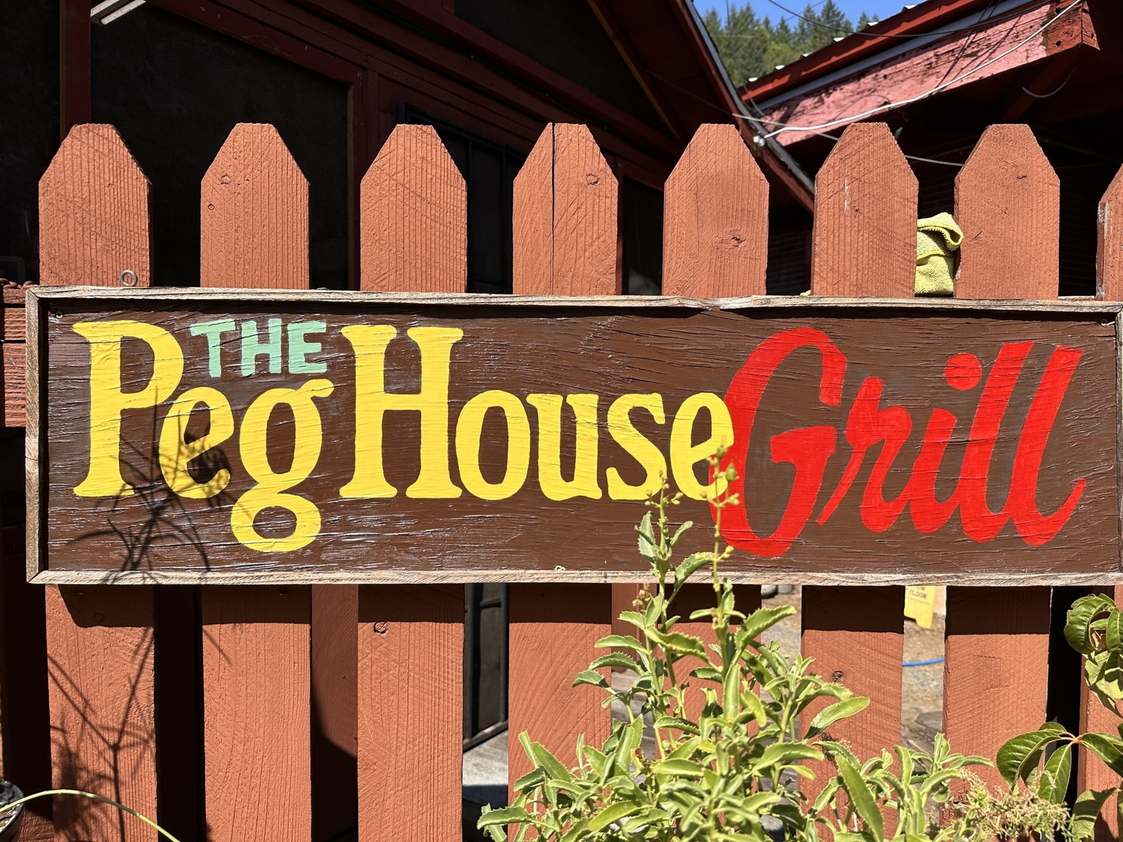 The image shows a wooden sign with &quot;The Peg House Grill&quot; painted in green, yellow, and red letters, mounted on a brown picket fence with some green plants in front.
