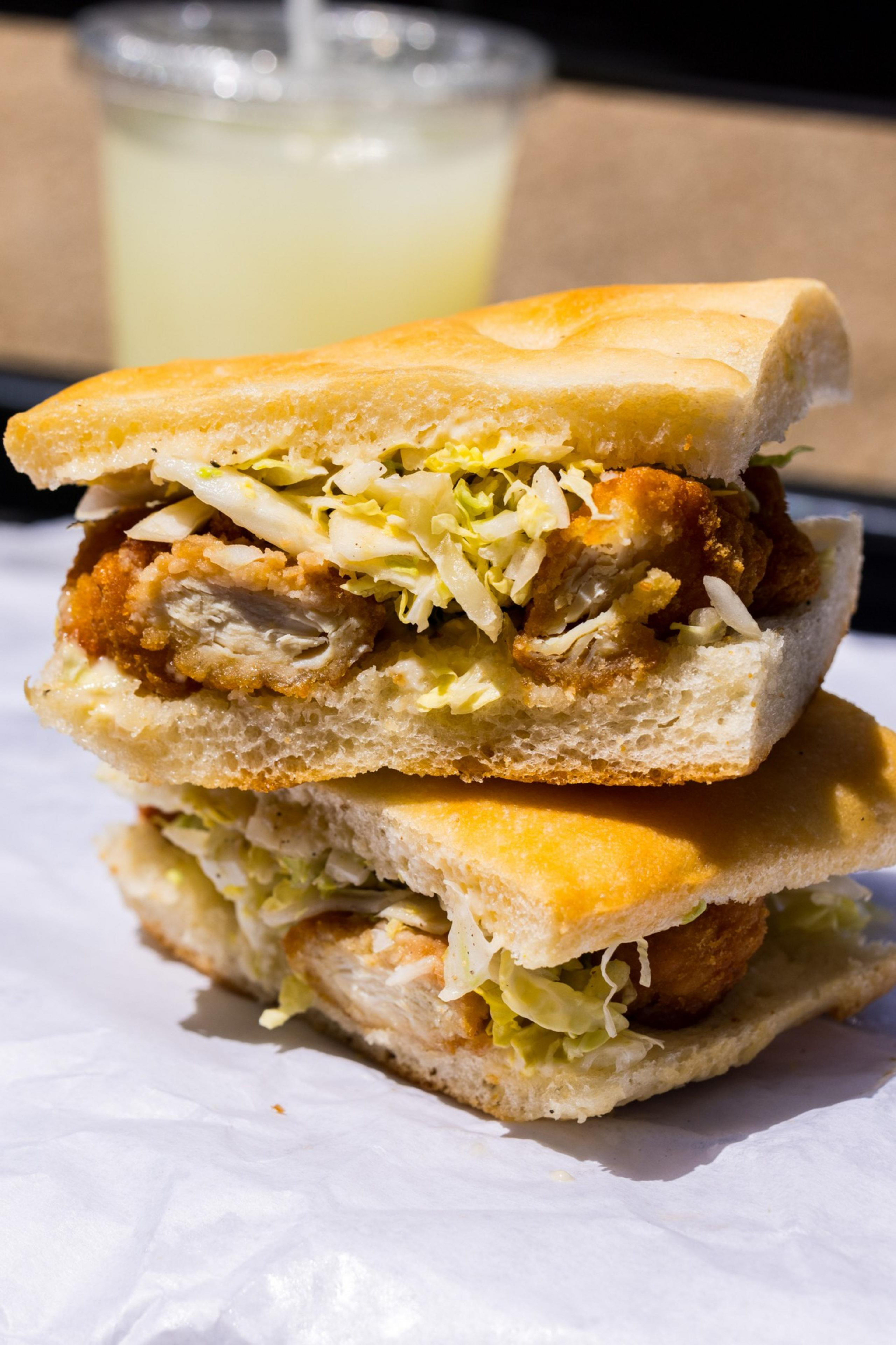A toasted sandwich with crispy chicken, shredded lettuce, and sauce sits on white paper with a cup of lemonade in the background.
