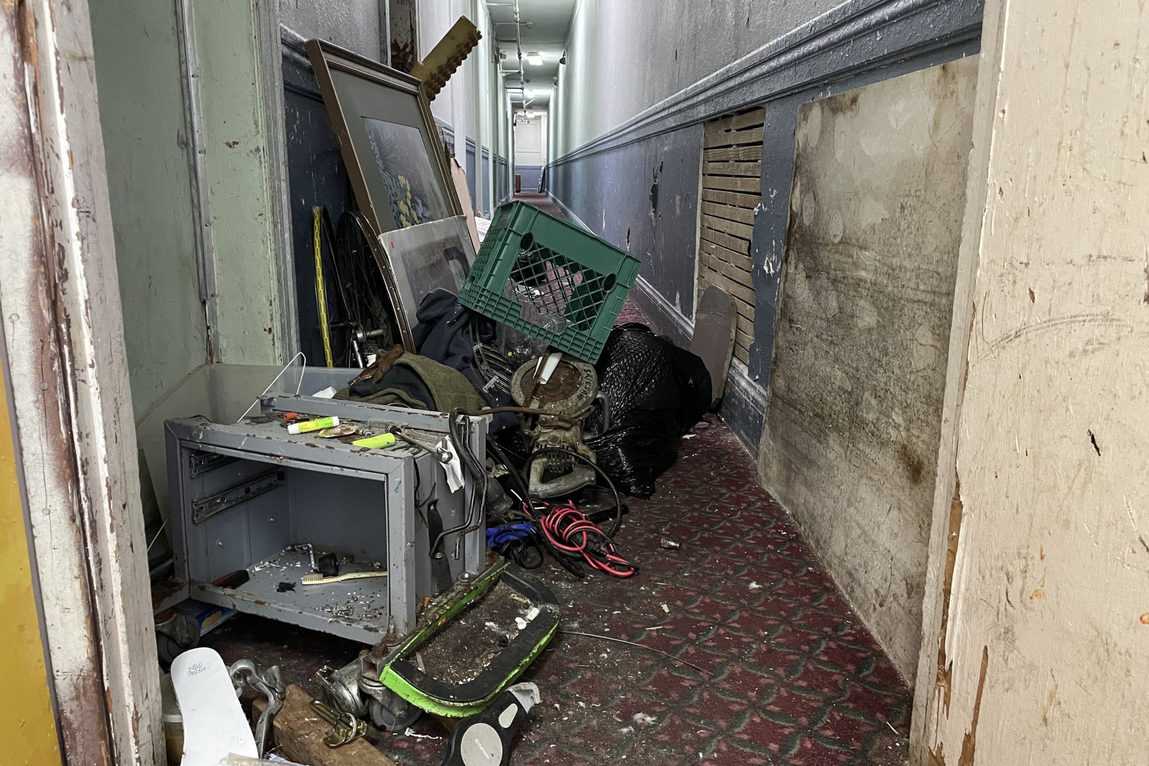 A cluttered hallway contains a mix of discarded items, including a broken TV, picture frame, bicycle wheel, and various debris, leading to a partially open door.