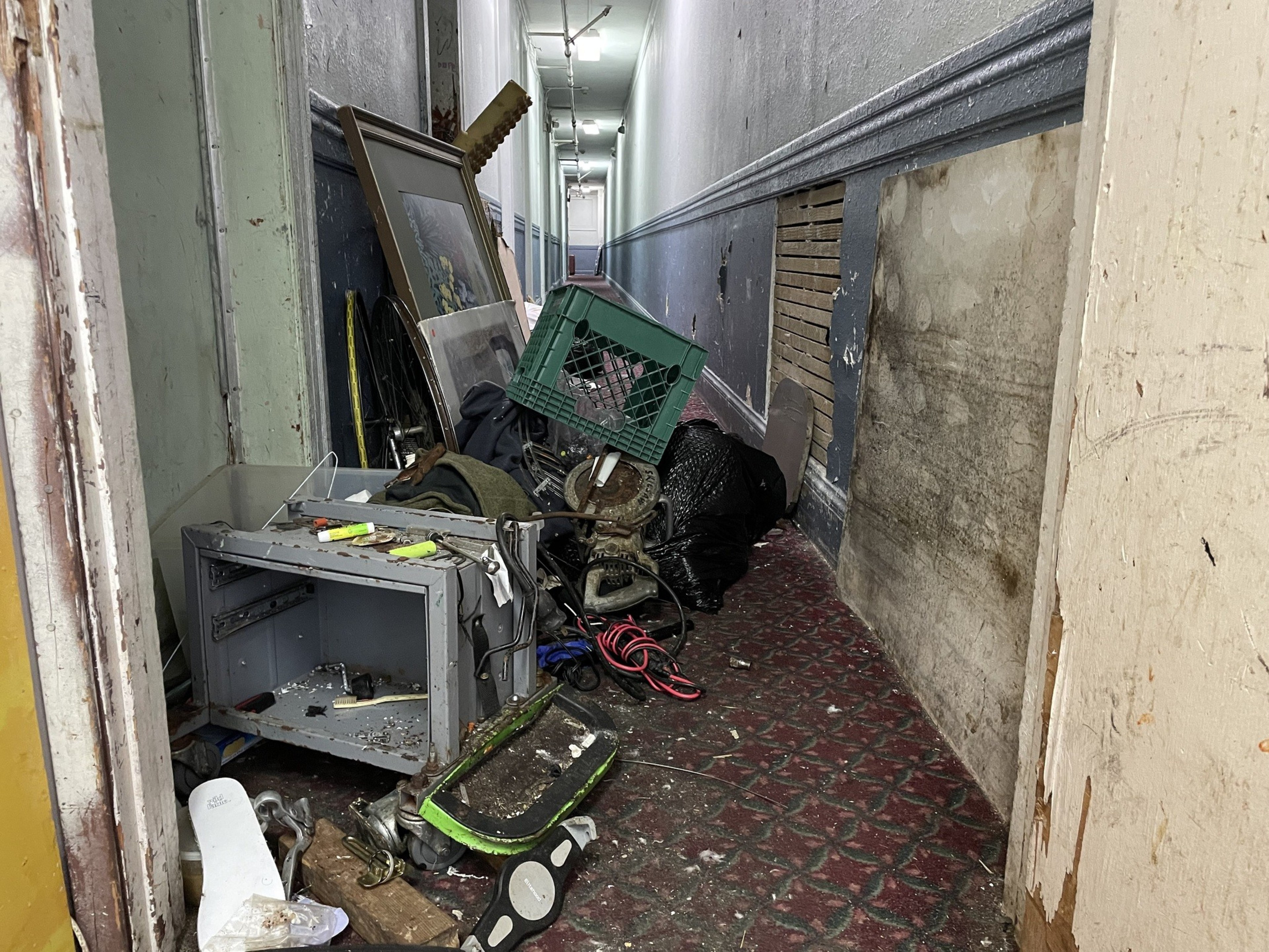 A cluttered hallway contains a mix of discarded items, including a broken TV, picture frame, bicycle wheel, and various debris, leading to a partially open door.