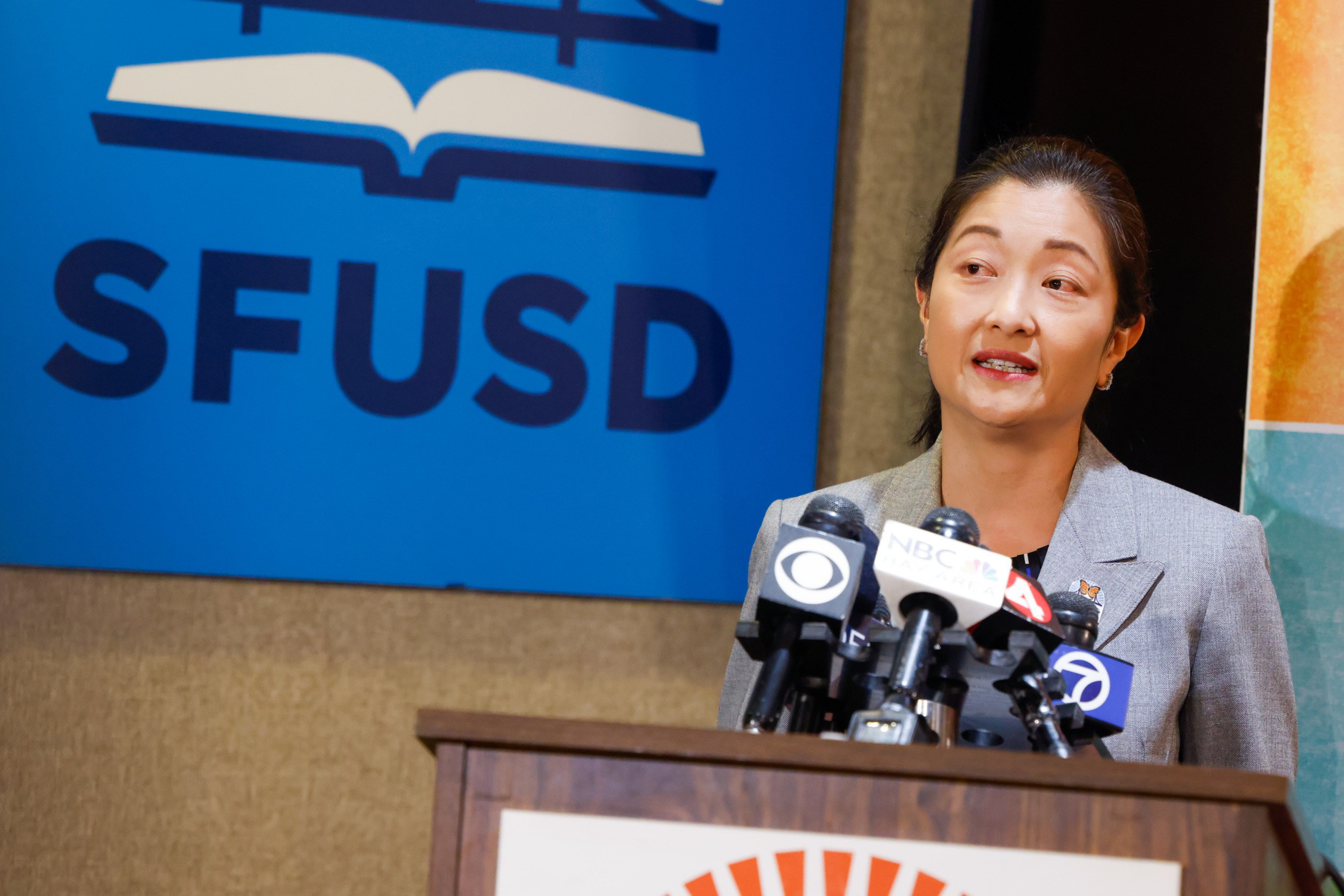 A woman in a suit speaks at a podium with multiple microphones. Behind her, a blue SFUSD sign with an open book logo is visible.