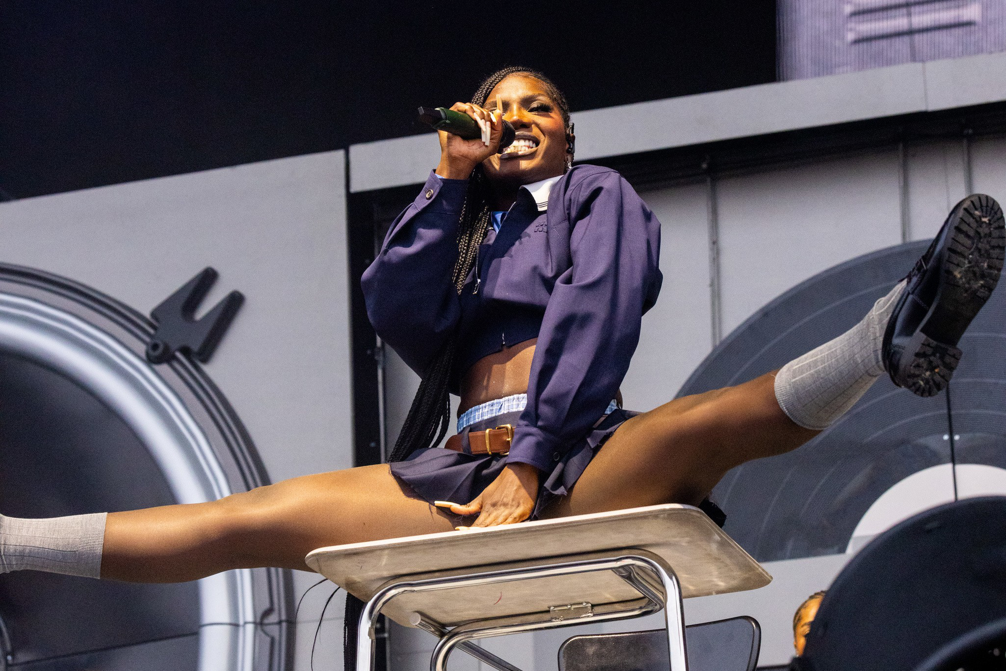 A female performer in a navy outfit does a full split on a school desk while singing into a microphone on stage.