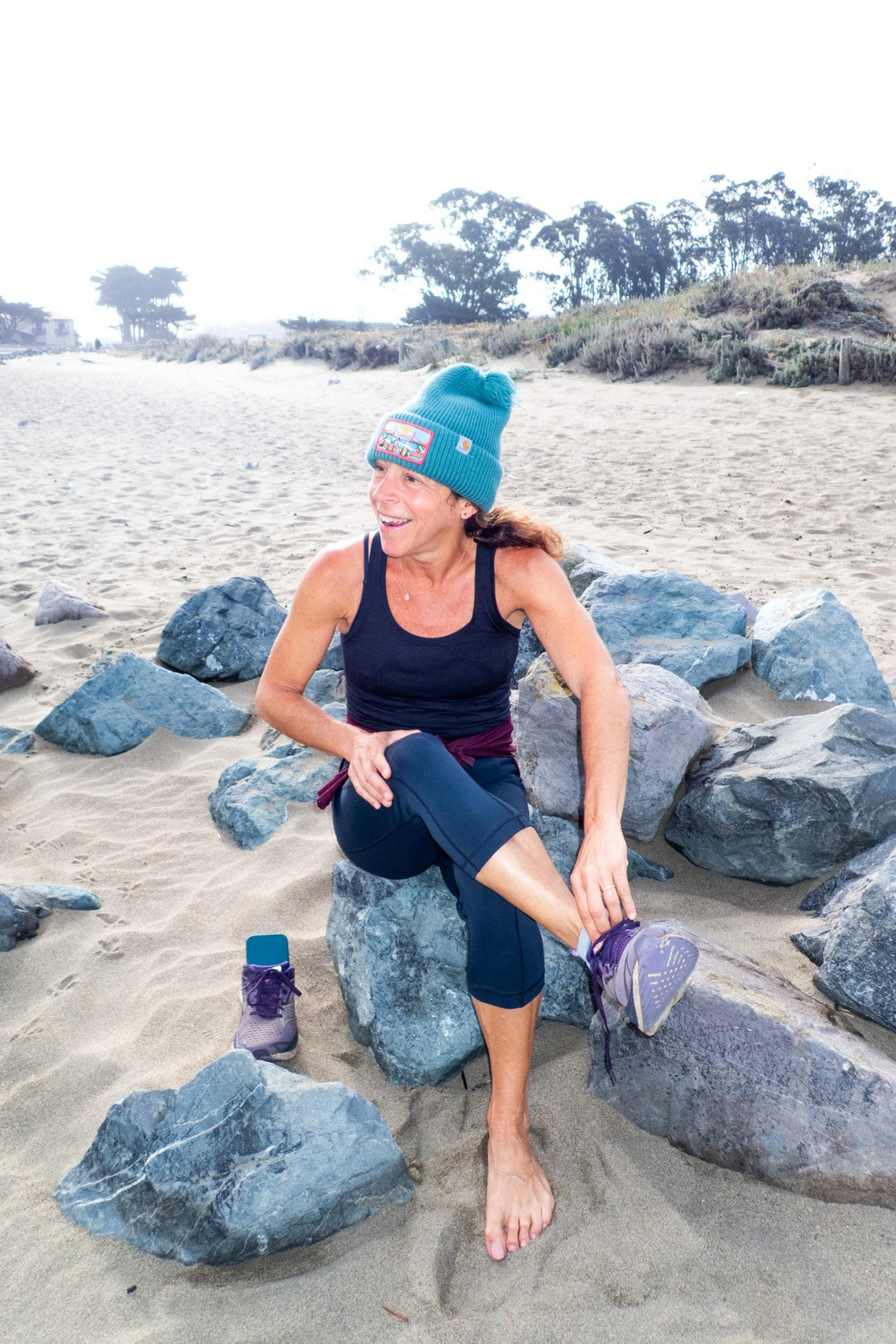 A woman in a turquoise beanie and black tank top sits on rocks at a beach, smiling as she ties her shoe. One shoe is off, and the beach is misty with trees in the background.